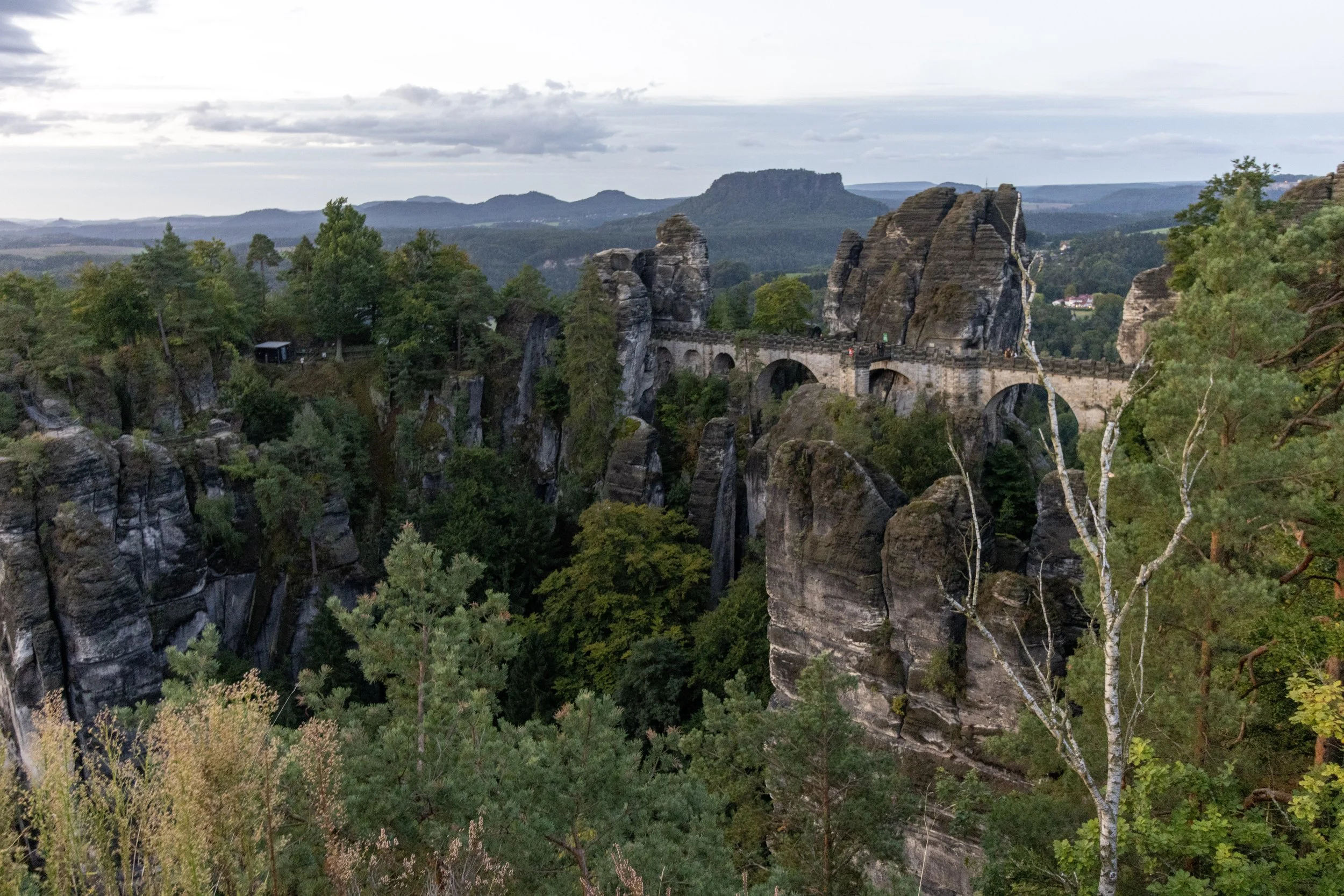 A stone bridge built on large rock formations over a deep forested valley, with rolling hills and distant mountains under a cloudy sky.
