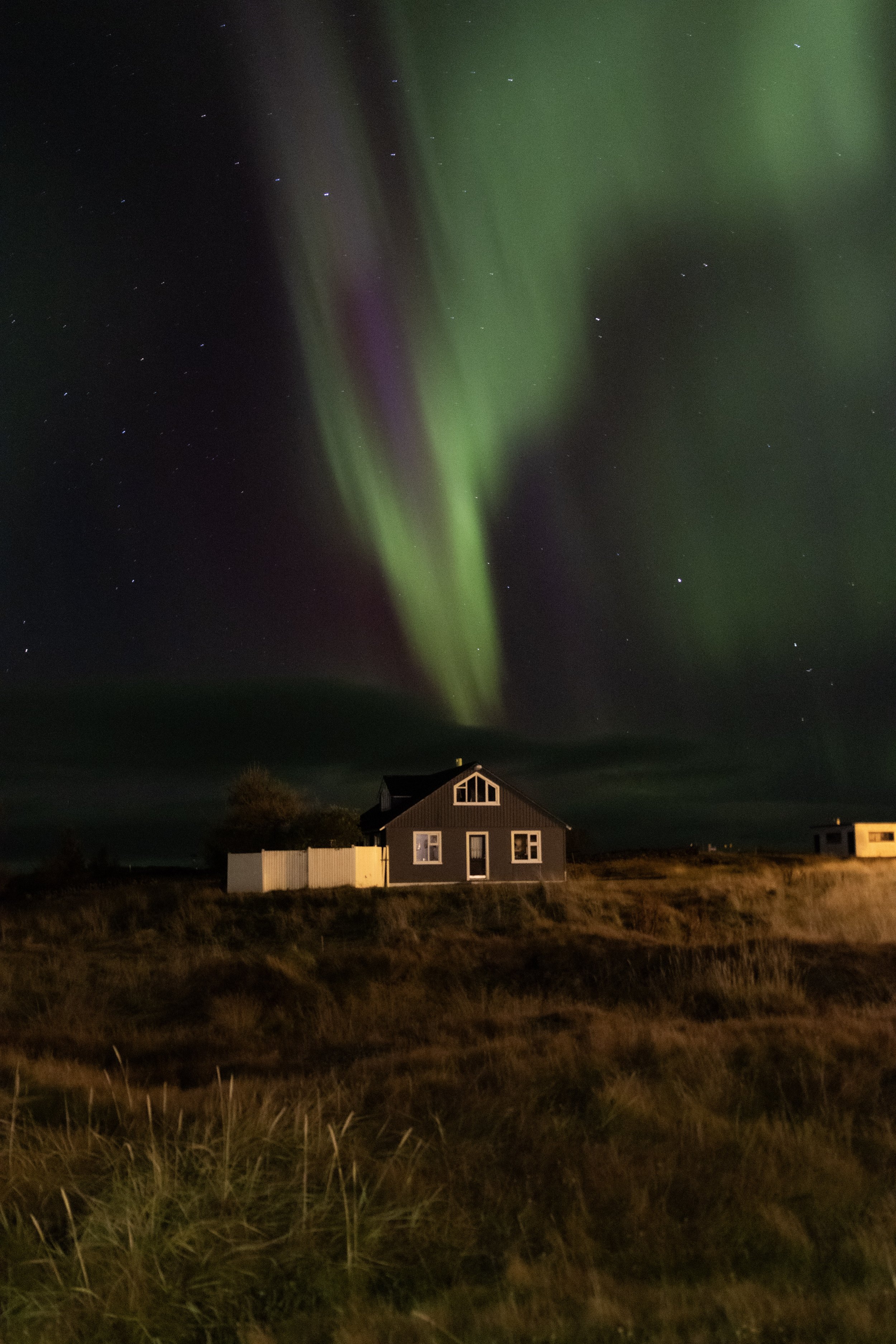 Night sky with vibrant northern lights over a house and grassy landscape.