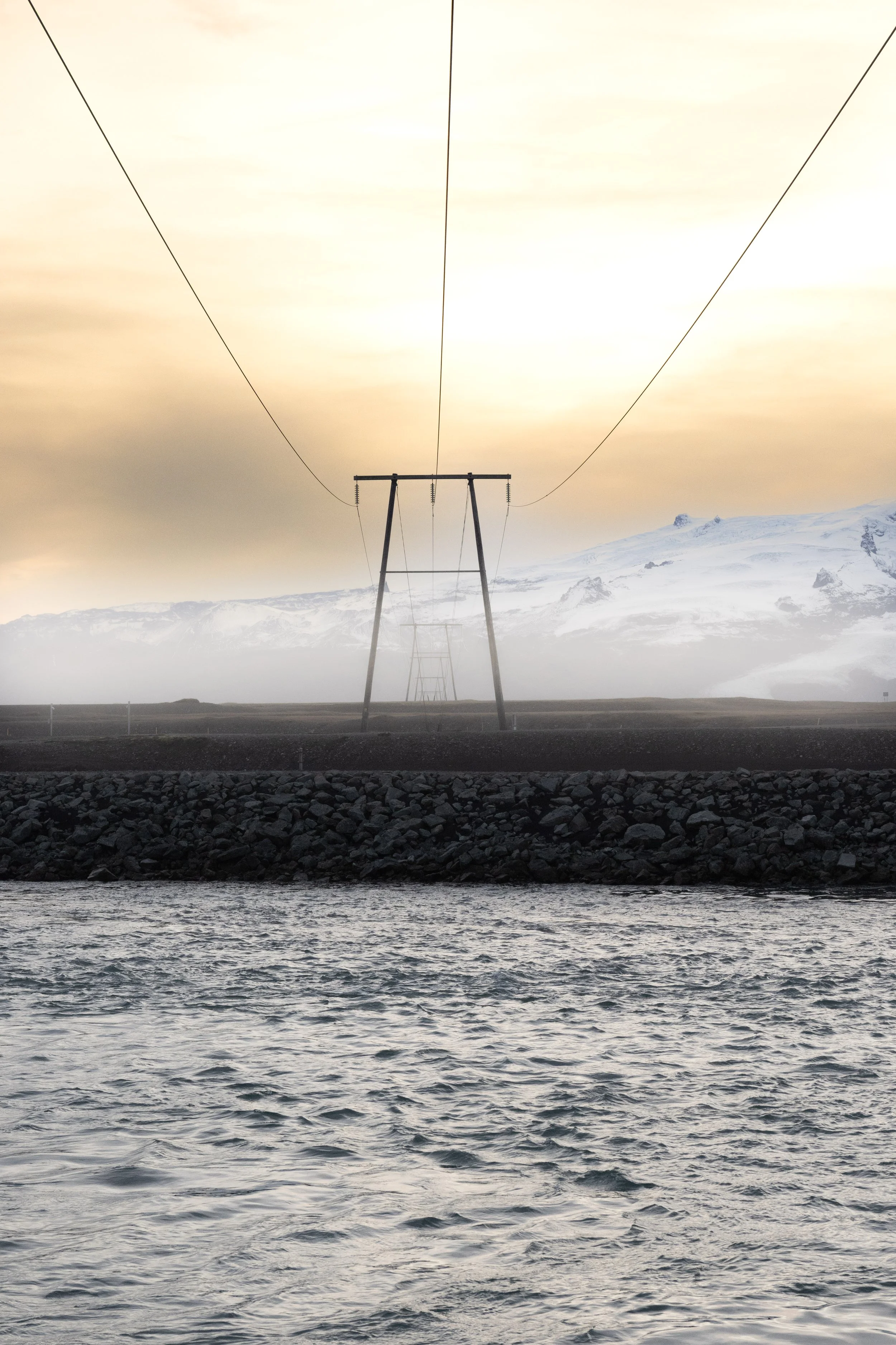 Power lines run over a body of water with a rocky shoreline, distant snow-capped mountains, and a hazy sky with warm tones.