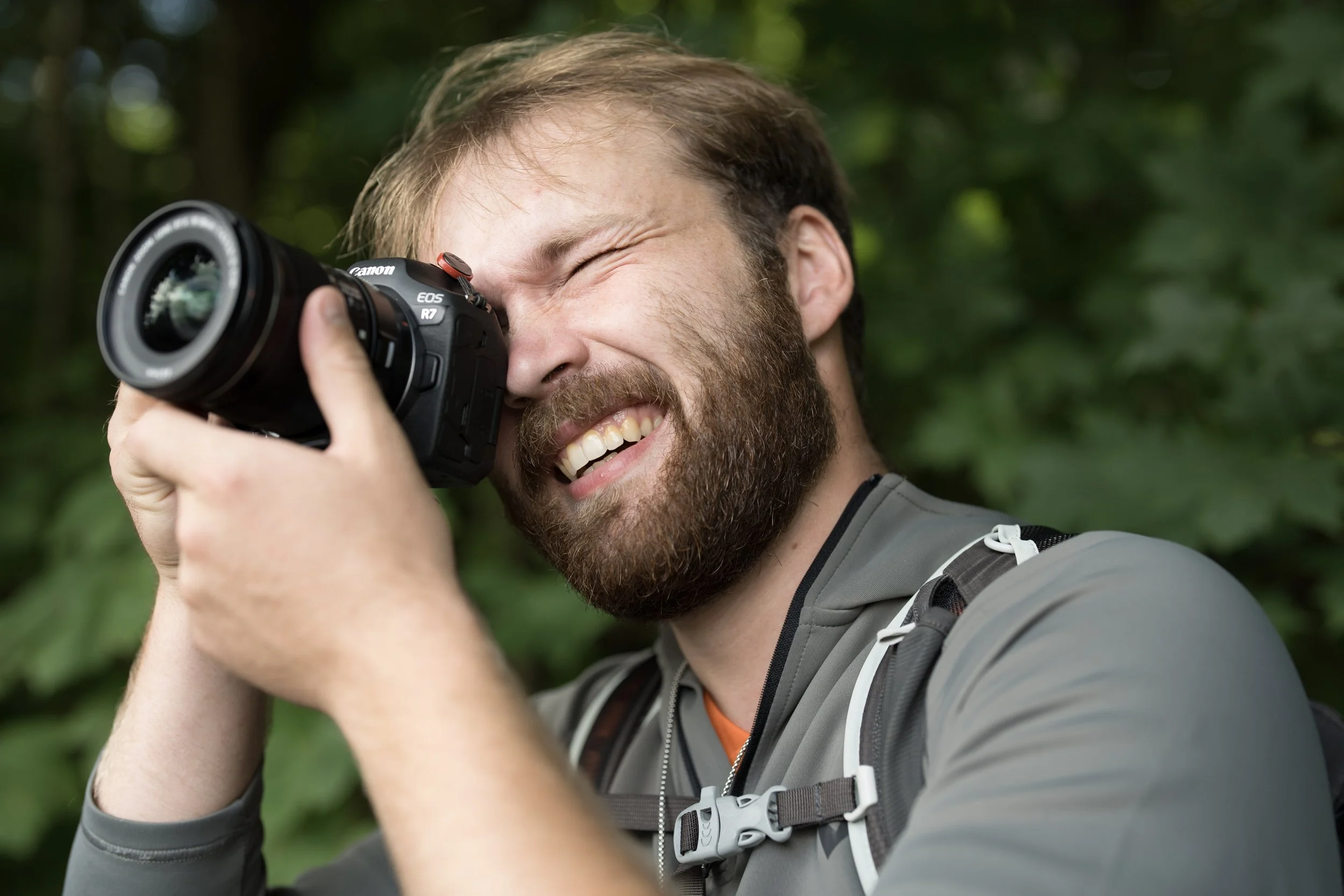 Man with a beard and gray jacket smiling while taking a photo outdoors with a camera.