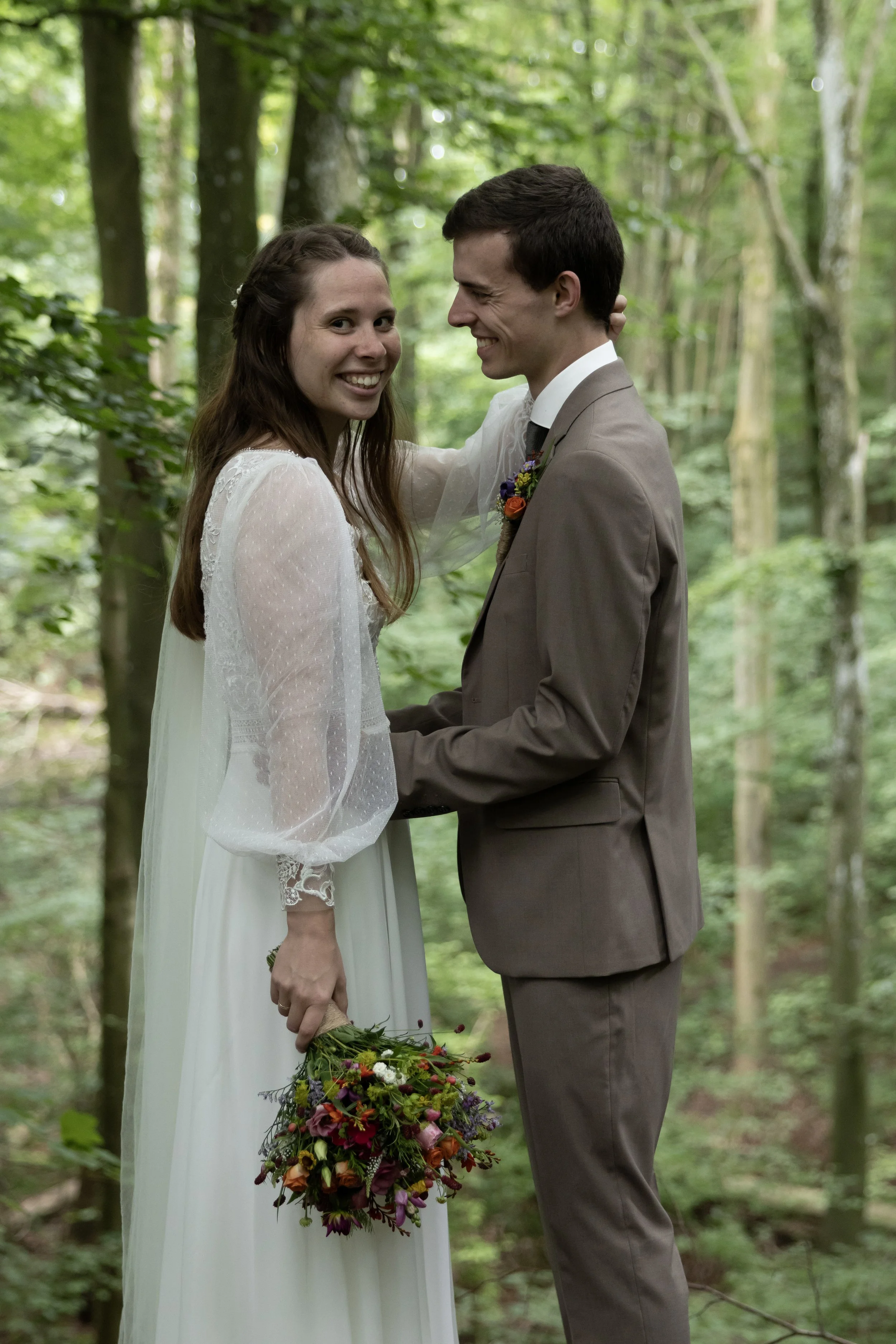 A bride and groom holding hands and smiling in a forest, the bride holding a bouquet of colorful flowers.