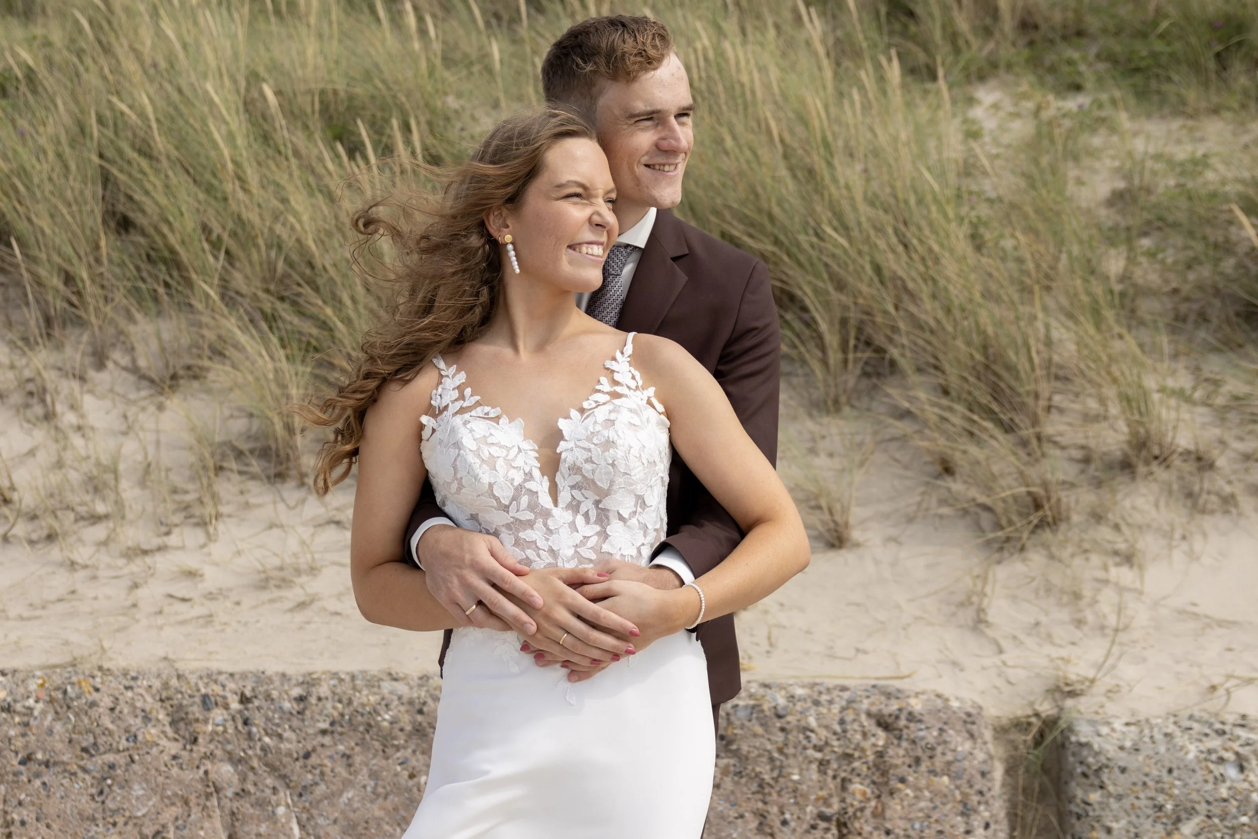 A newlywed couple in wedding attire standing on a sandy beach with tall grasses in the background, smiling and embracing each other.
