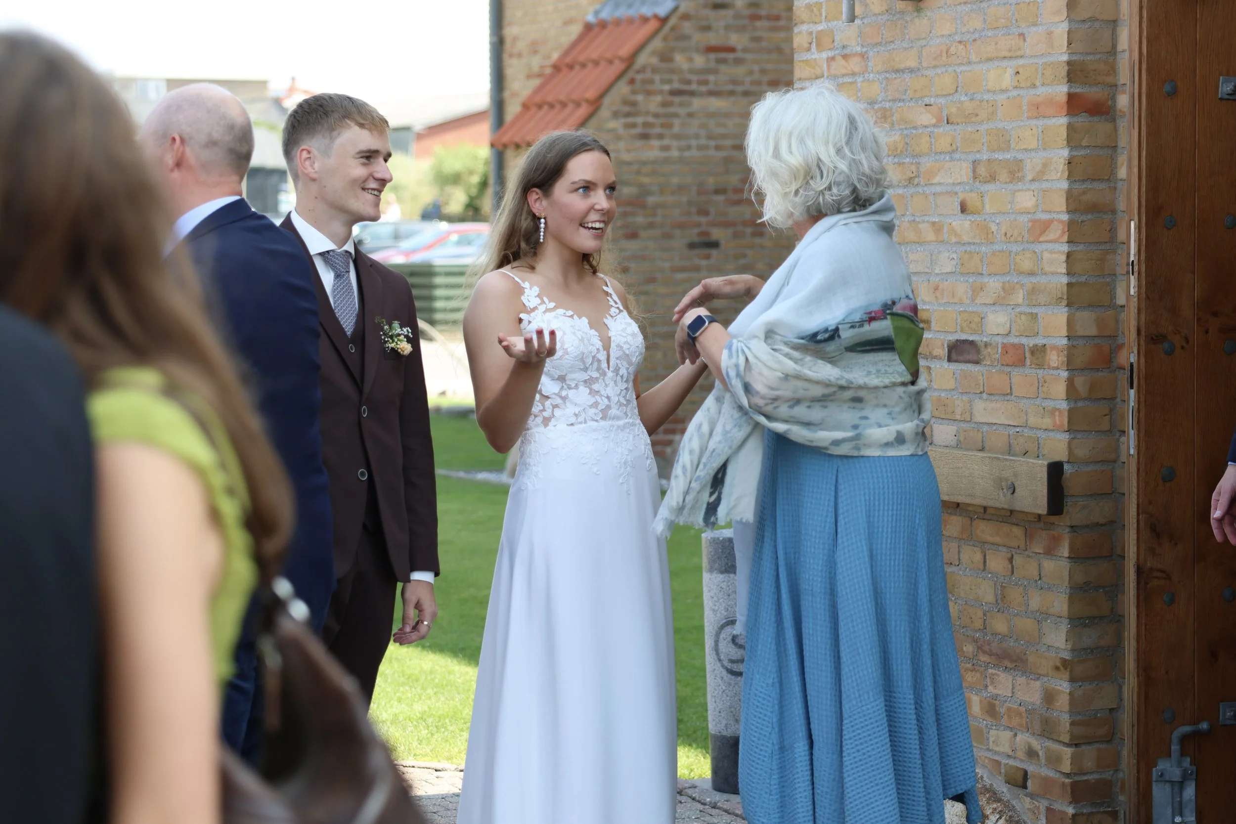 A bride in a white wedding dress talking to an older woman with gray hair, who is wearing a blue skirt and a light shawl, at an outdoor wedding reception.