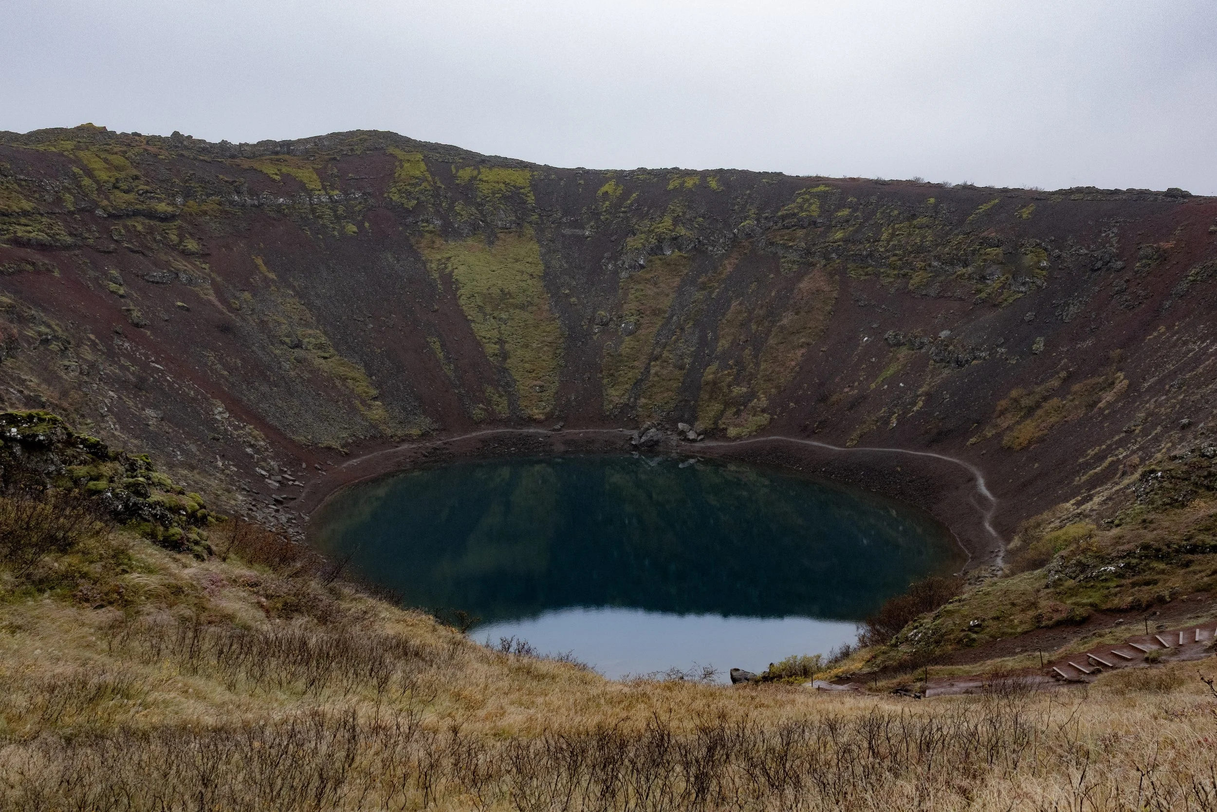 A crater lake inside a volcanic caldera with steep rocky walls covered in green moss and vegetation, seen during overcast weather.
