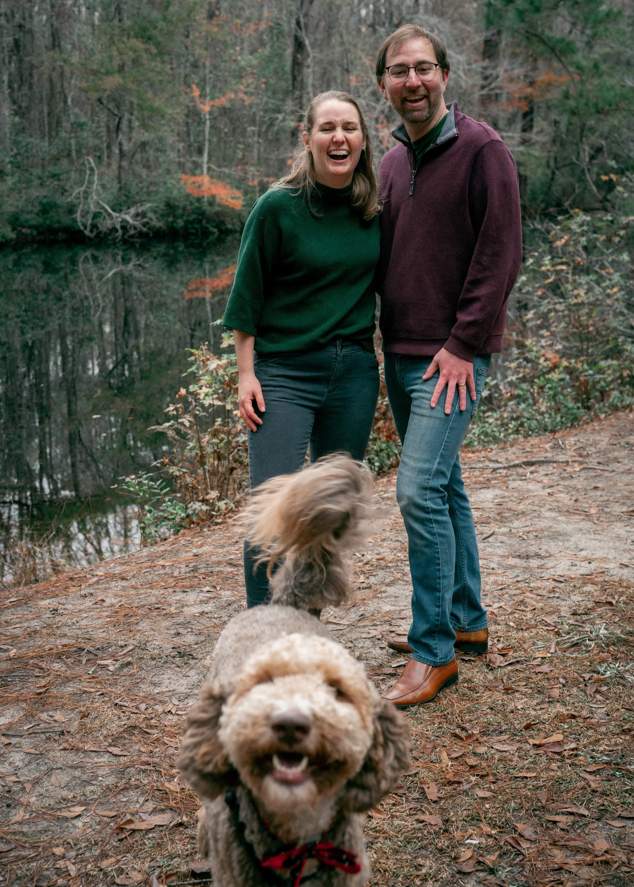 Julia and Dan laughing outdoors on a wooded trail near a lake, with a smiling Moose in the foreground.