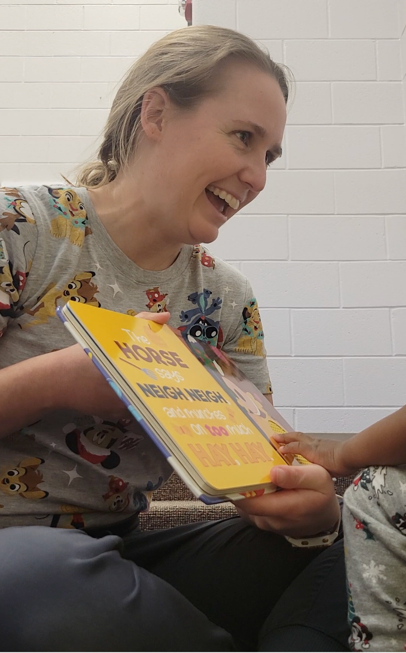 A woman with blonde hair smiling and holding a book titled 'The Horse Says Neigh Neigh' while a child's hand reaches toward the book.