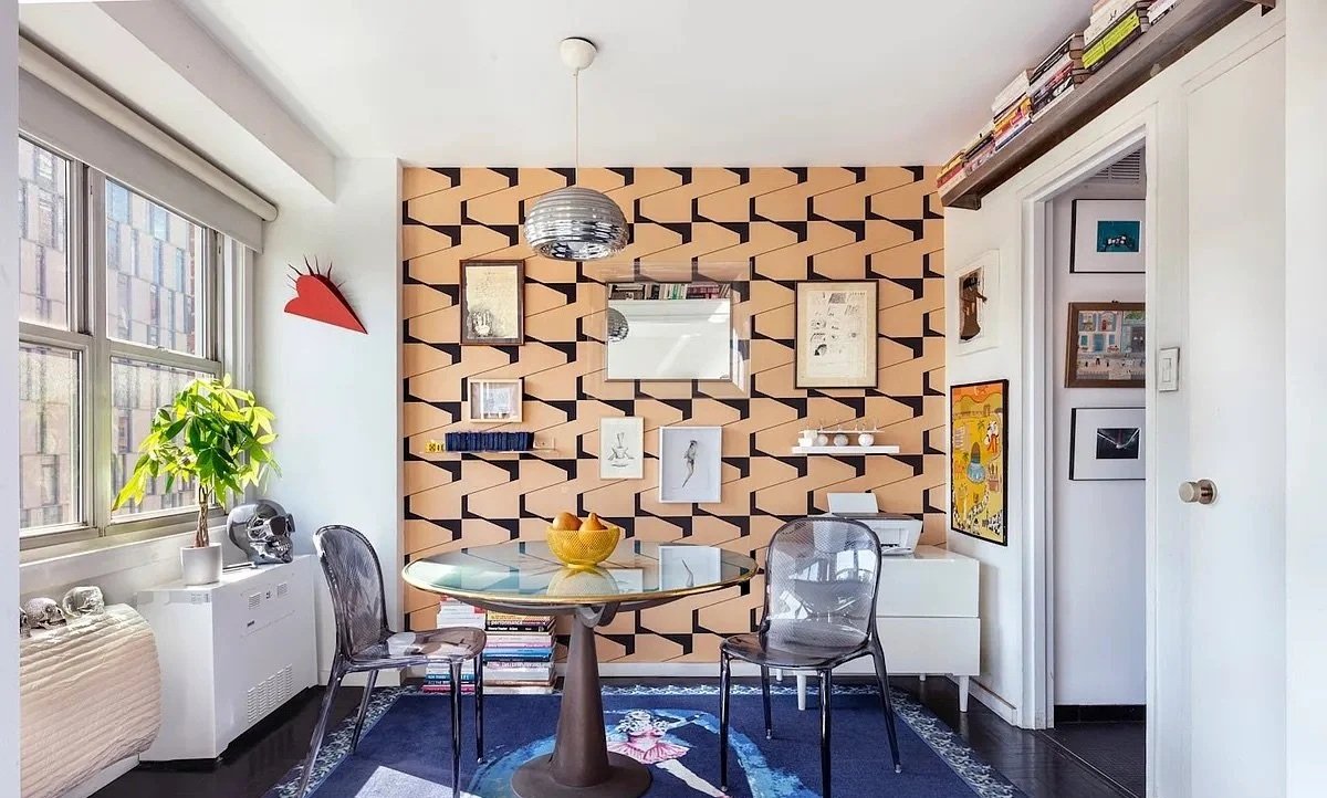 An image of a dining room with colorful wallpaper and hanging wall art. There is a table in the center of the room, and two translucent chairs.