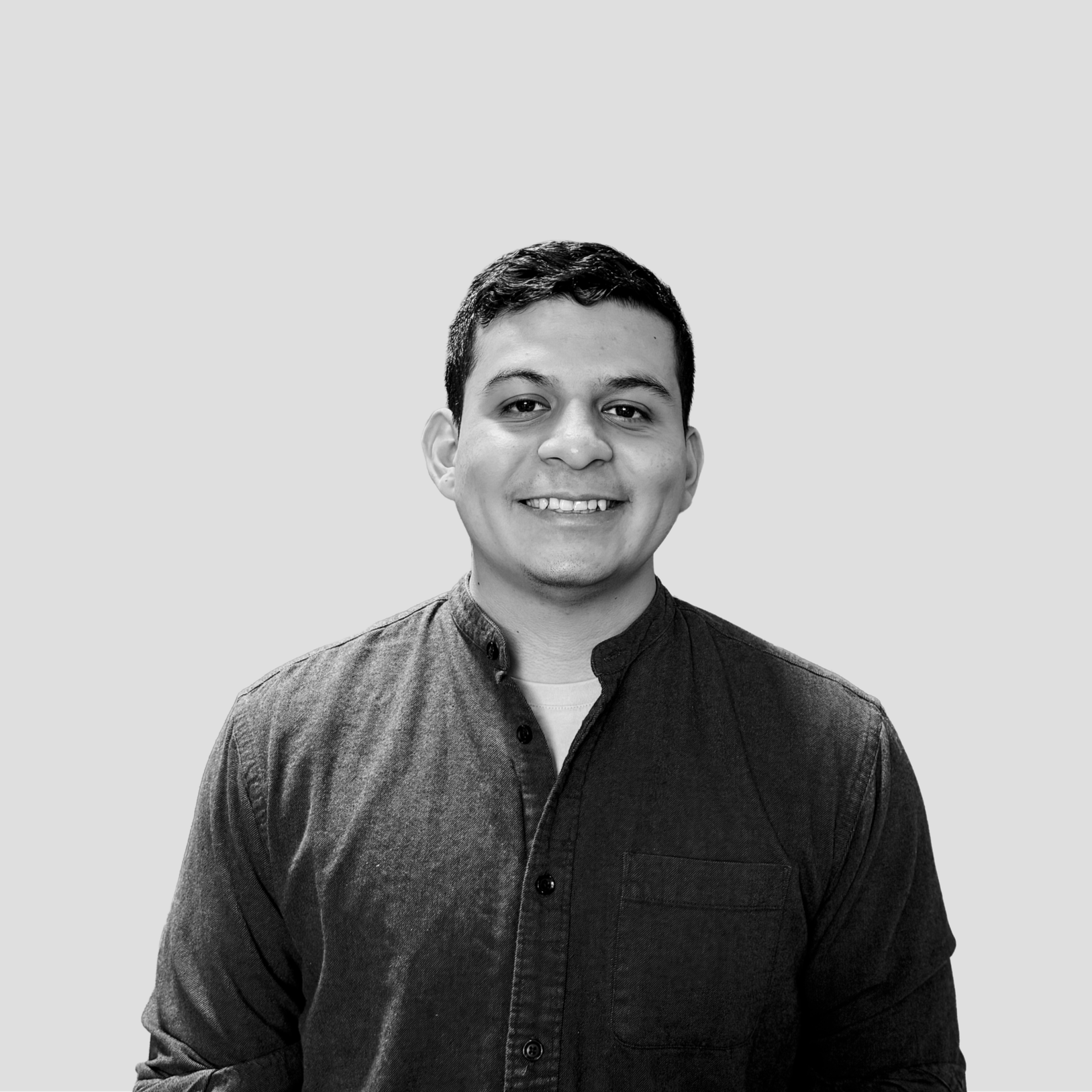 Black and white portrait of a smiling young man with short dark hair, wearing a dark button-up shirt with a mandarin collar, against a plain light gray background.