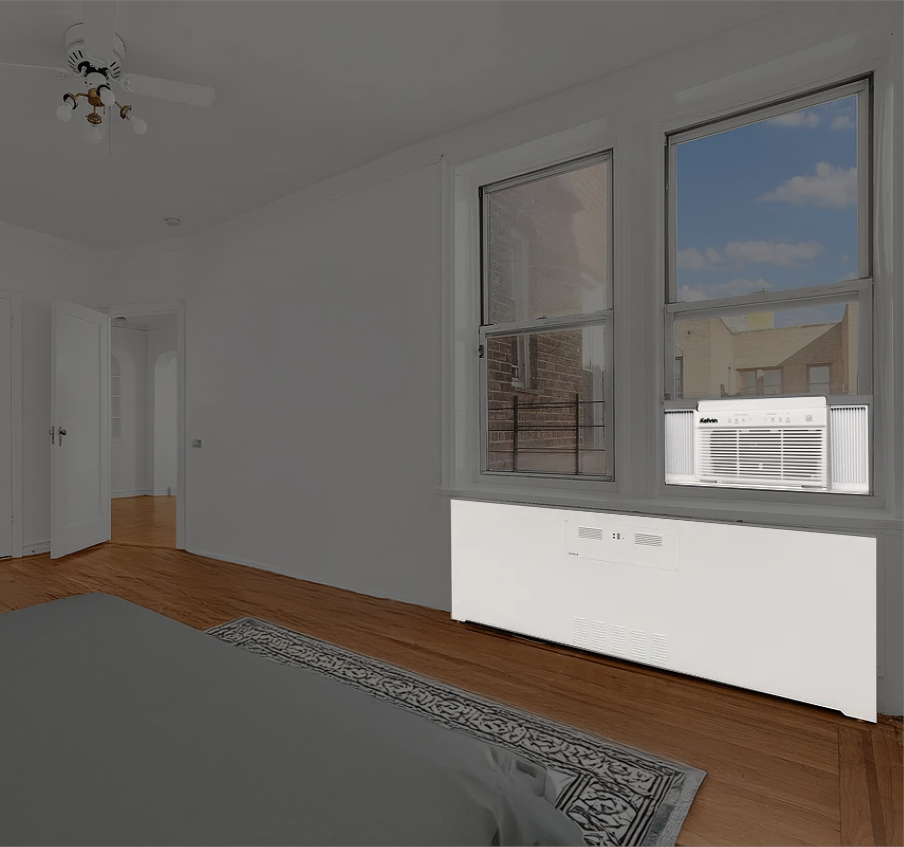 A bright, empty bedroom with white walls, a wooden floor, and a ceiling fan. There are two large windows, one with an air conditioning unit installed. An open door leads to another room.