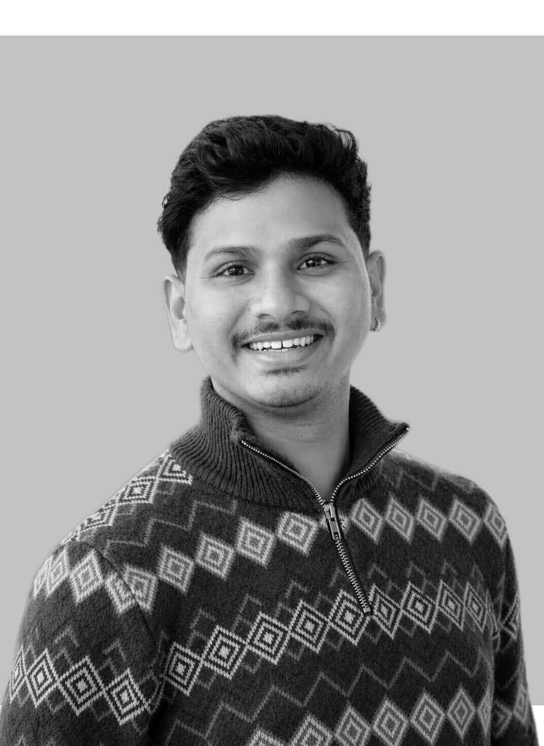 A young man with short, curly hair looking at the camera, wearing a textured sweater over a long-sleeve shirt, against a plain light background.