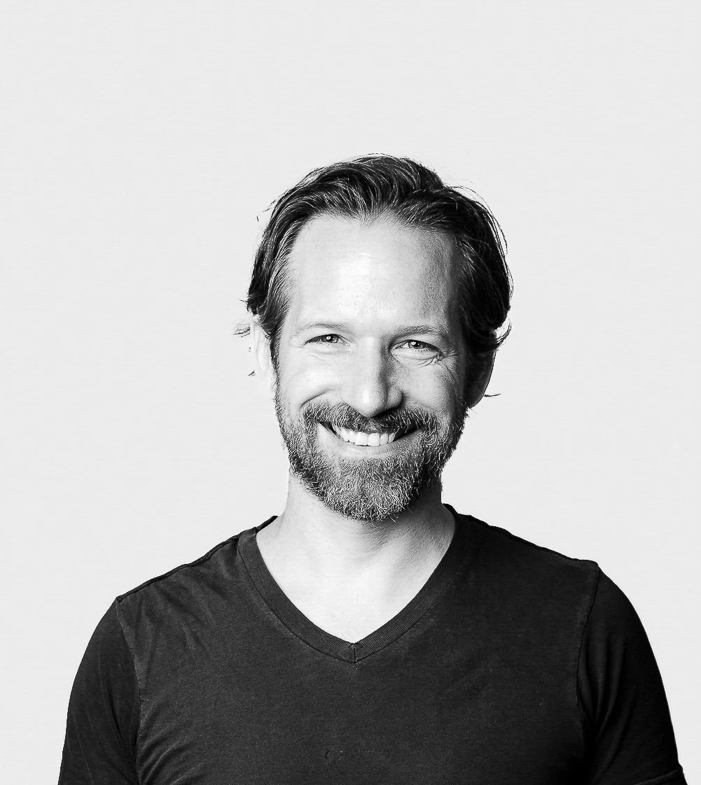Black and white portrait of a smiling man with a beard and mustache, wearing a dark V-neck shirt, against a plain light background.