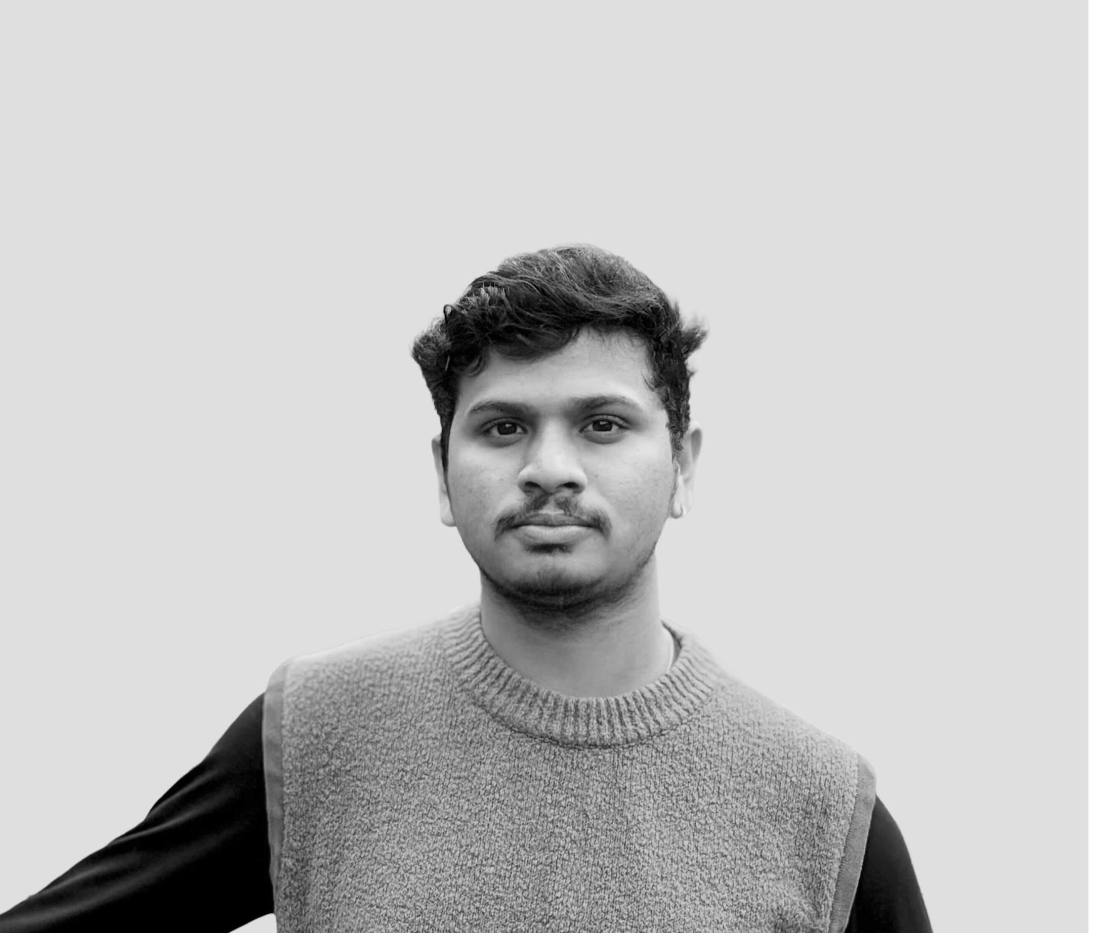 A young man with short, curly hair looking at the camera, wearing a textured sweater over a long-sleeve shirt, against a plain light background.