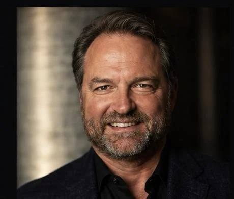 A smiling man with a beard, wearing a suit jacket and dress shirt, against a plain white background.