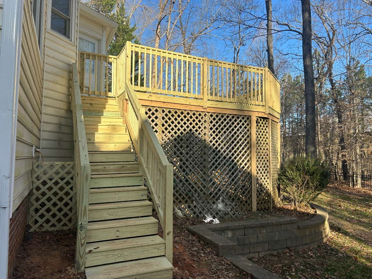 Newly built wooden deck with stairs attached to a house, with lattice skirting underneath, surrounded by trees and a small garden bed.