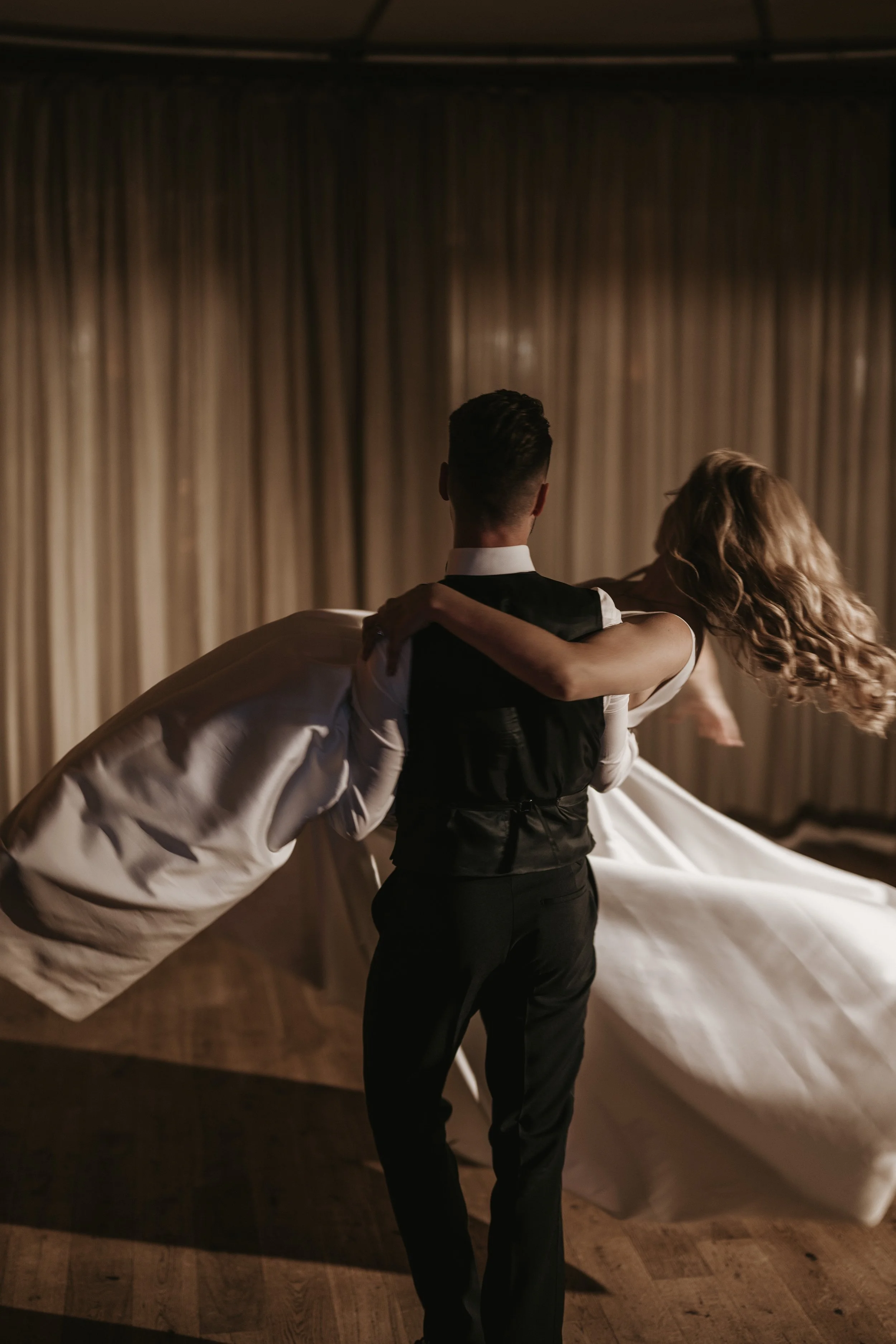 A man in a tuxedo is dipping a woman in a white dress for a dance in a dimly lit room with beige curtains.