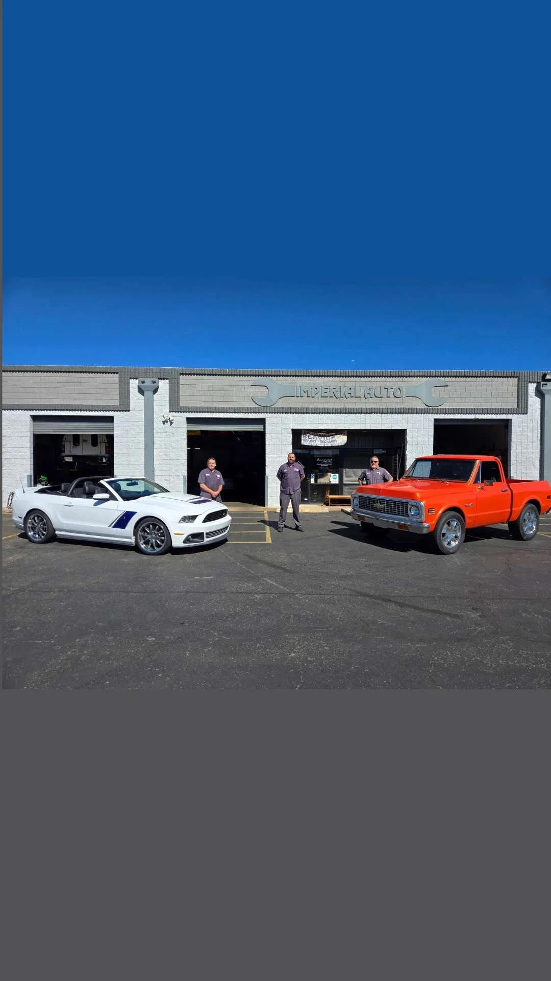 Front view of an auto repair shop with a white convertible sports car and an orange pickup truck parked outside, and three employees standing in front.