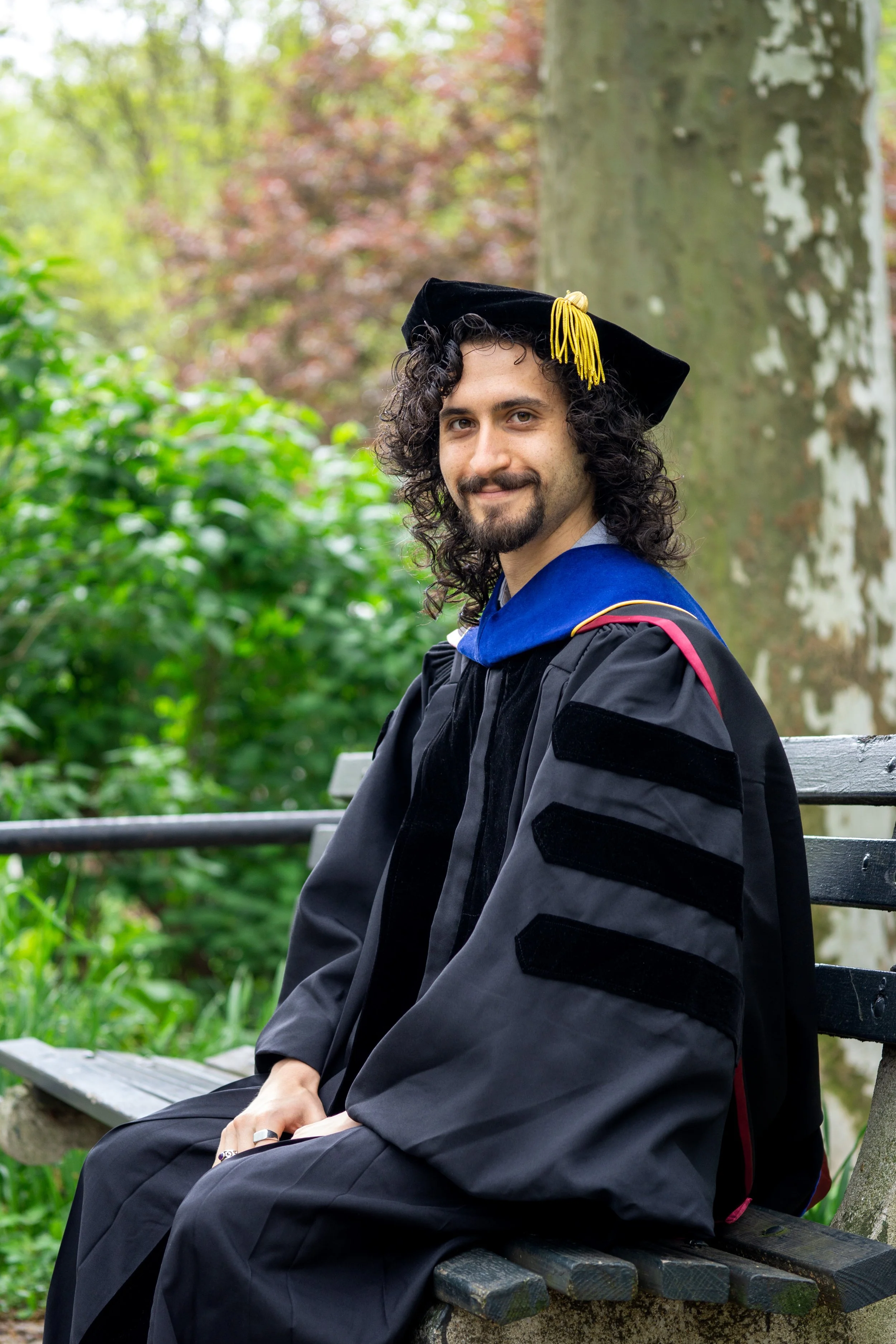 A man wearing academic regalia and a graduation cap with tassel sitting on a park bench outdoors, smiling.