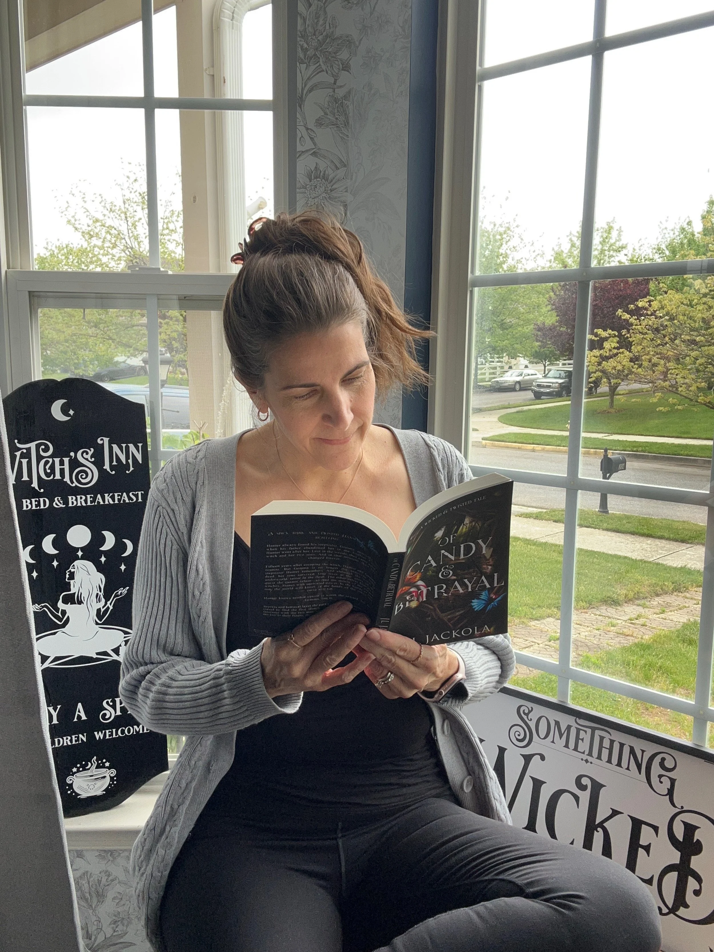 Author J L Jackola sitting by a large window reading a book titled 'Of Candy & Betrayal' with decorative signs on the windowsill.