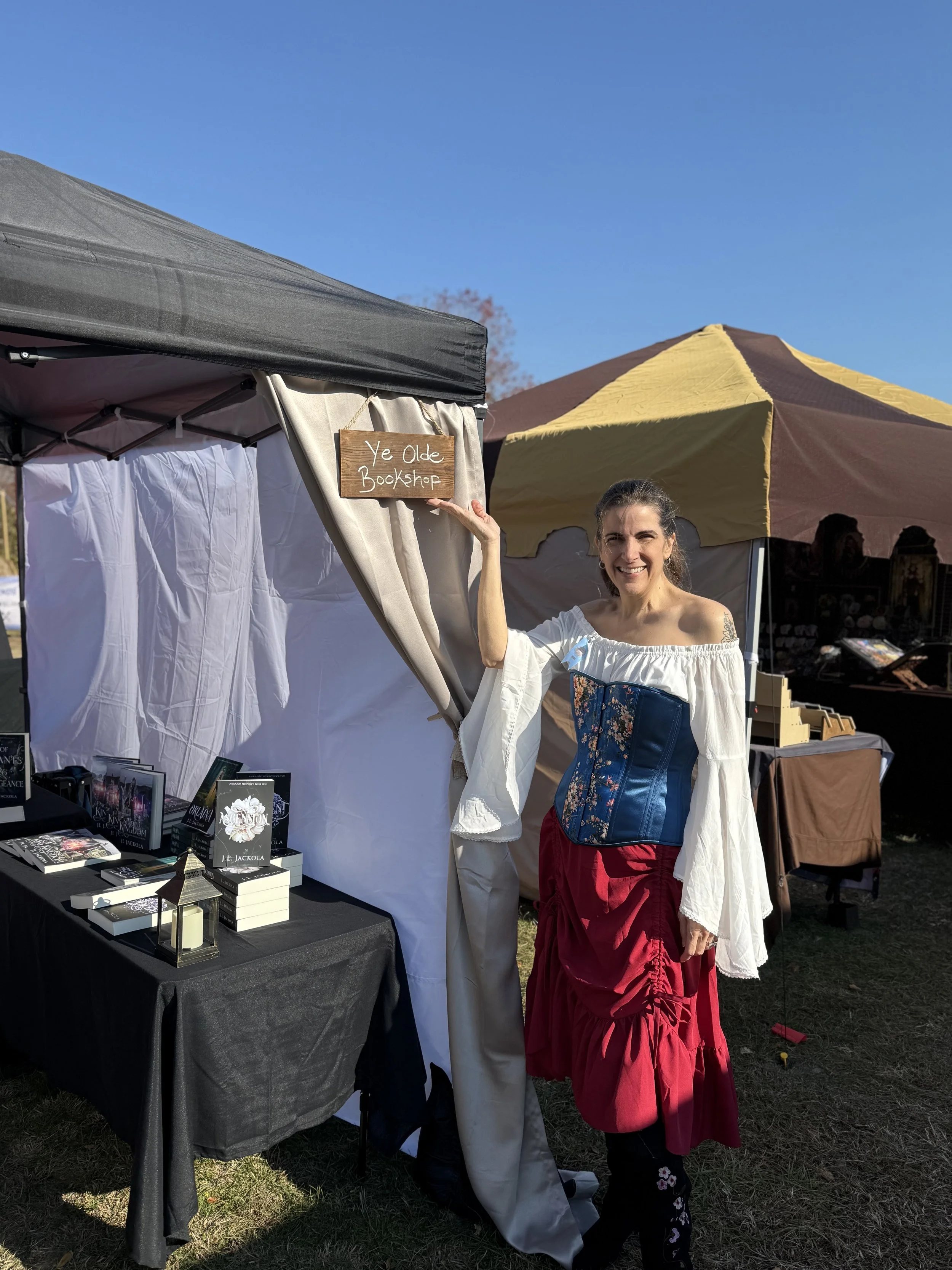 Author J L Jackola dressed in a white off-shoulder blouse, a blue corset, and a red layered skirt standing next to a booth at an outdoor market, holding a wooden sign that reads 'Ye Olde Bookshop'.