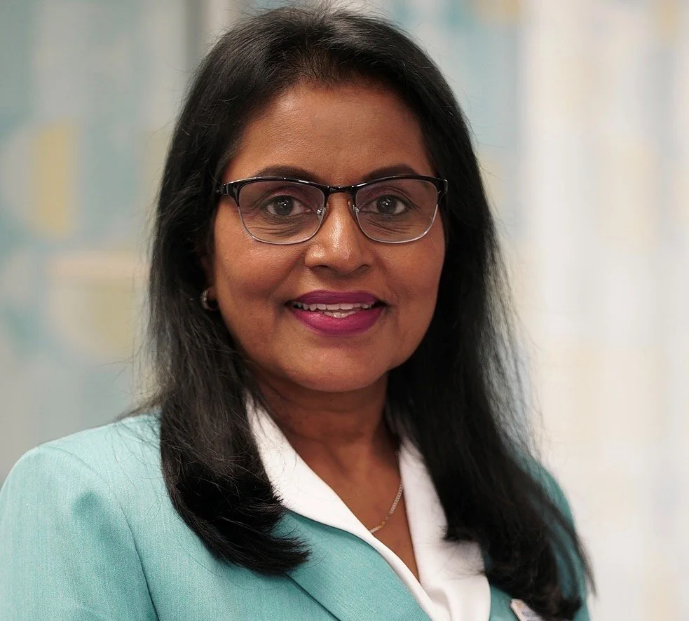A woman with black hair wearing glasses and a teal blazer, smiling, in front of a blurred background.