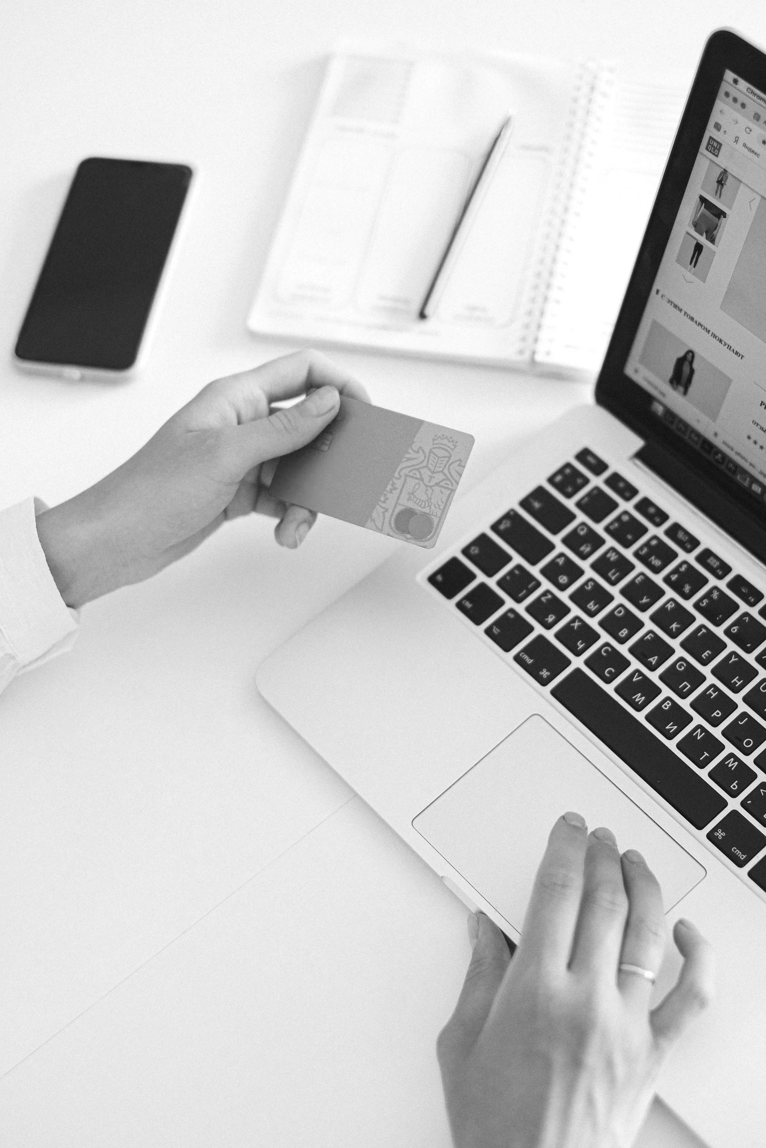 Person holding a credit card near a laptop and smartphone on a desk, with an open planner and pencil.