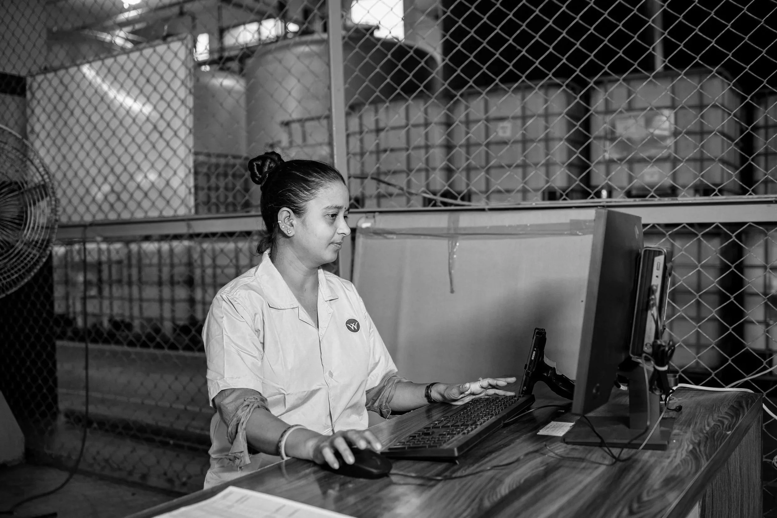 A woman using a computer at a desk in an industrial or warehouse setting, with large containers and a metal fence in the background.
