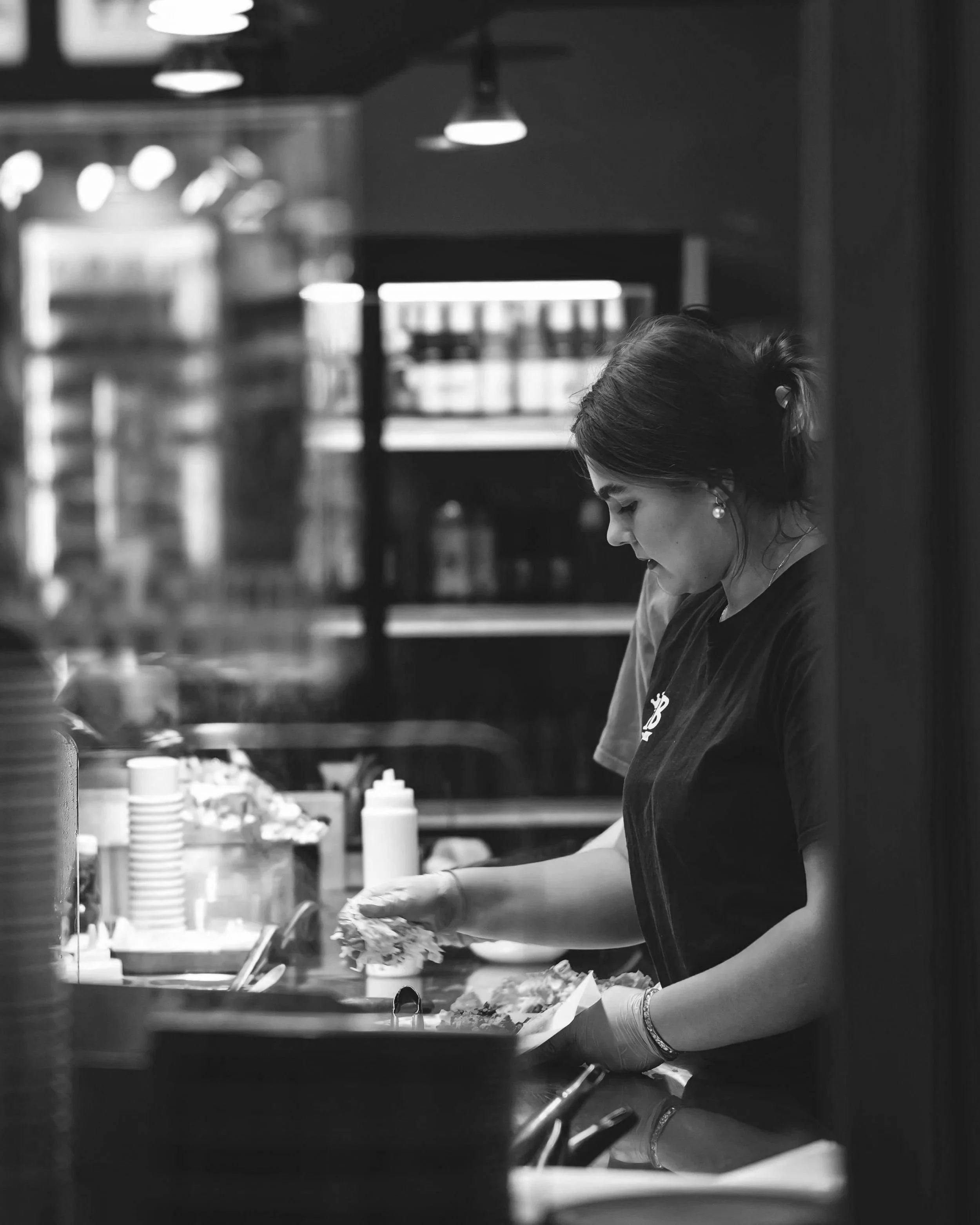 A woman wearing gloves and earrings working behind a counter in a kitchen, preparing food. The scene is in black and white, with shelves and kitchen supplies in the background.