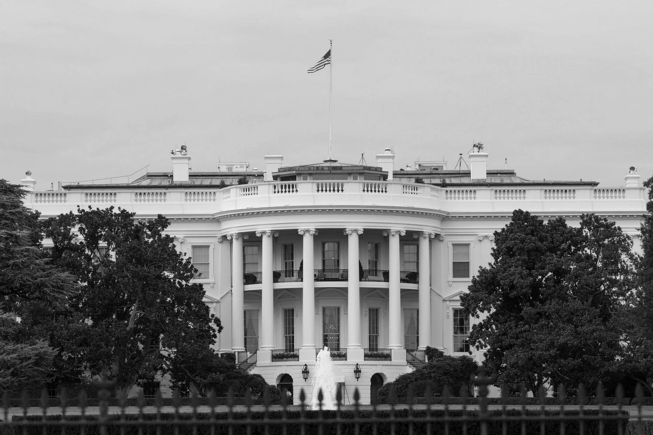 The White House, the official residence and workplace of the President of the United States, seen in this black and white photograph with trees surrounding it and a fountain in front.