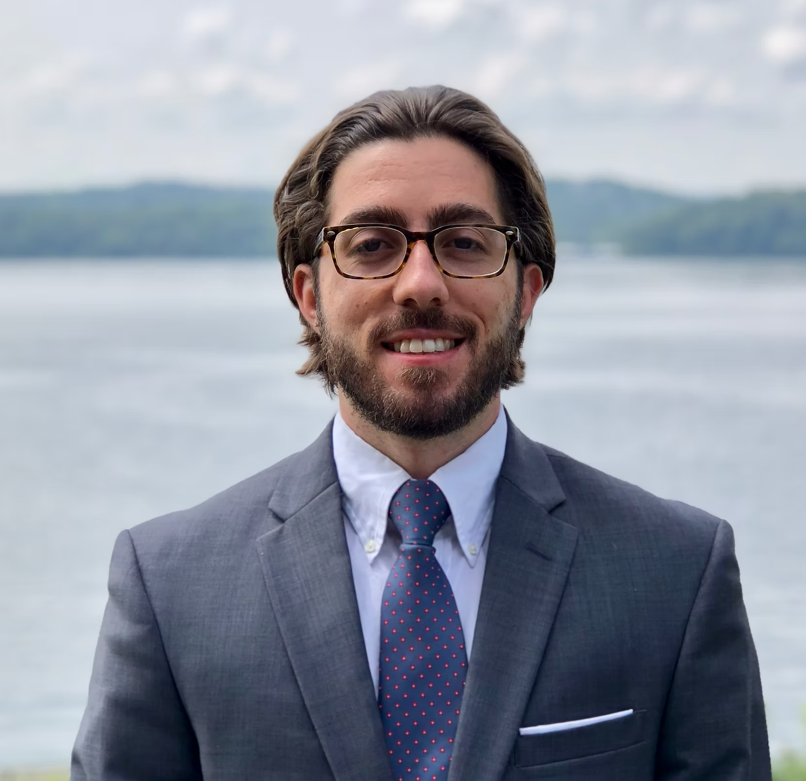 A man with dark hair, beard, and glasses wearing a gray suit, white shirt, and a patterned tie stands outdoors near a body of water with trees and hills in the background.
