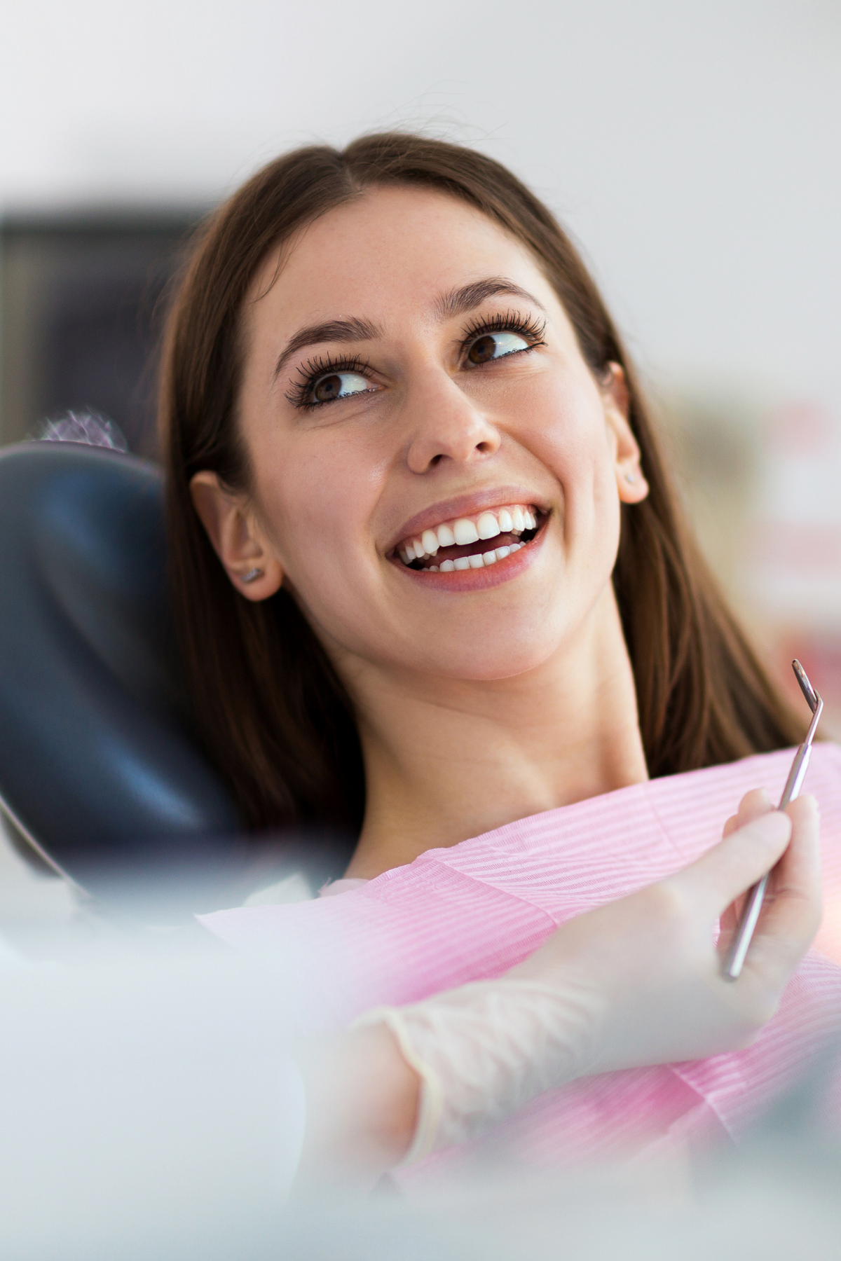 A young woman at the dentist's office, smiling and looking slightly to the side, wearing a pink dental gown and holding a dental tool.
