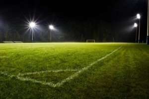 Campo de fútbol iluminado durante la noche con línea blanca y portería en fondo.