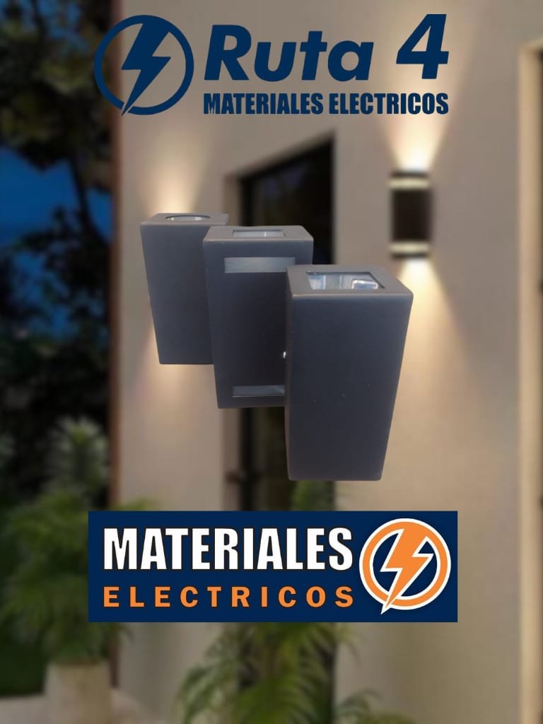Cartel publicitario de Materiales Eléctricos con texto en azul y naranja, y logotipo de un rayo dentro de un círculo, en un entorno exterior con plantas y la fachada de un edificio.