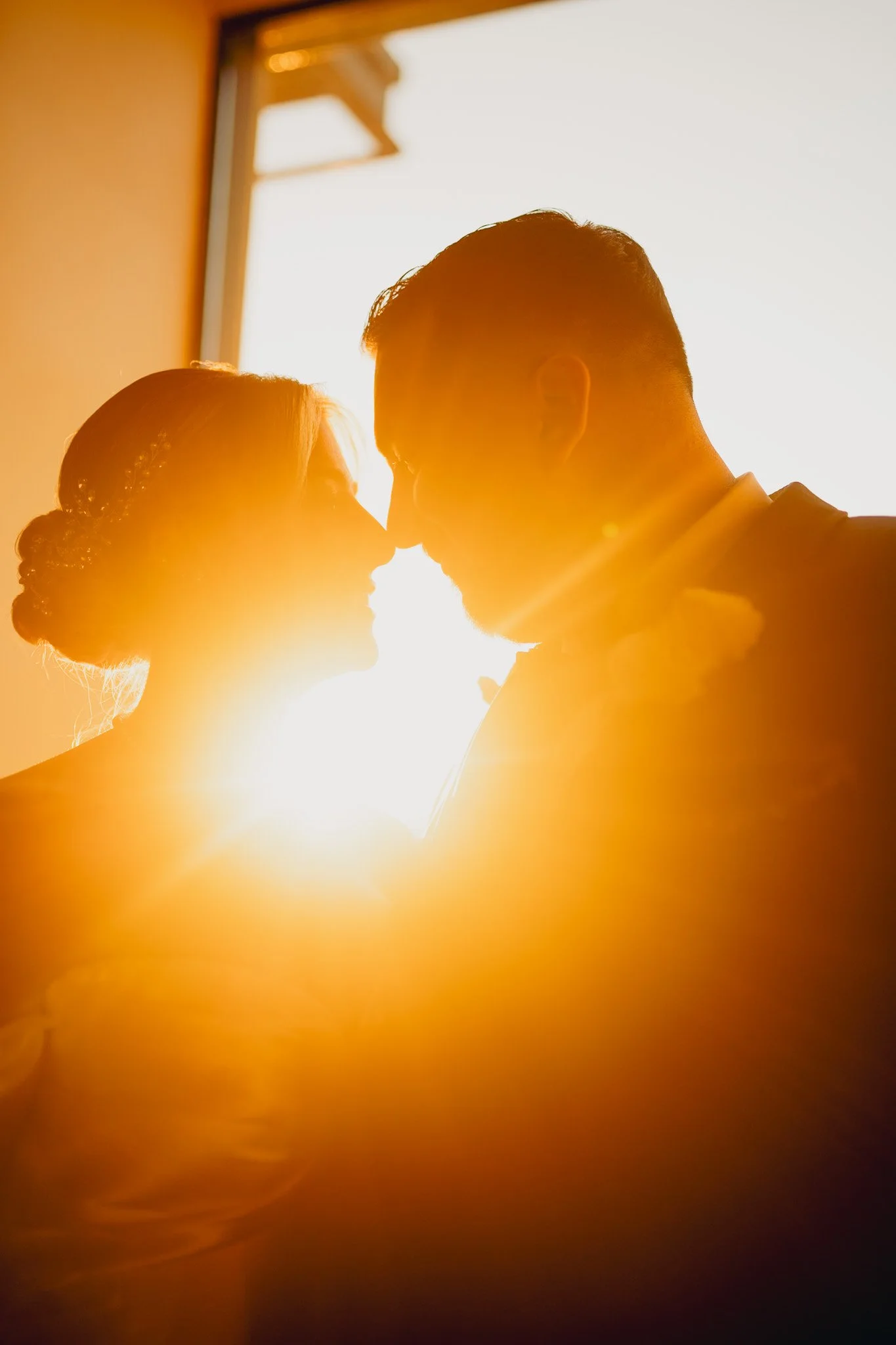 Silhouette of a bride and groom touching foreheads, backlit by warm sunlight, with the bride wearing a veil and the groom in a suit.