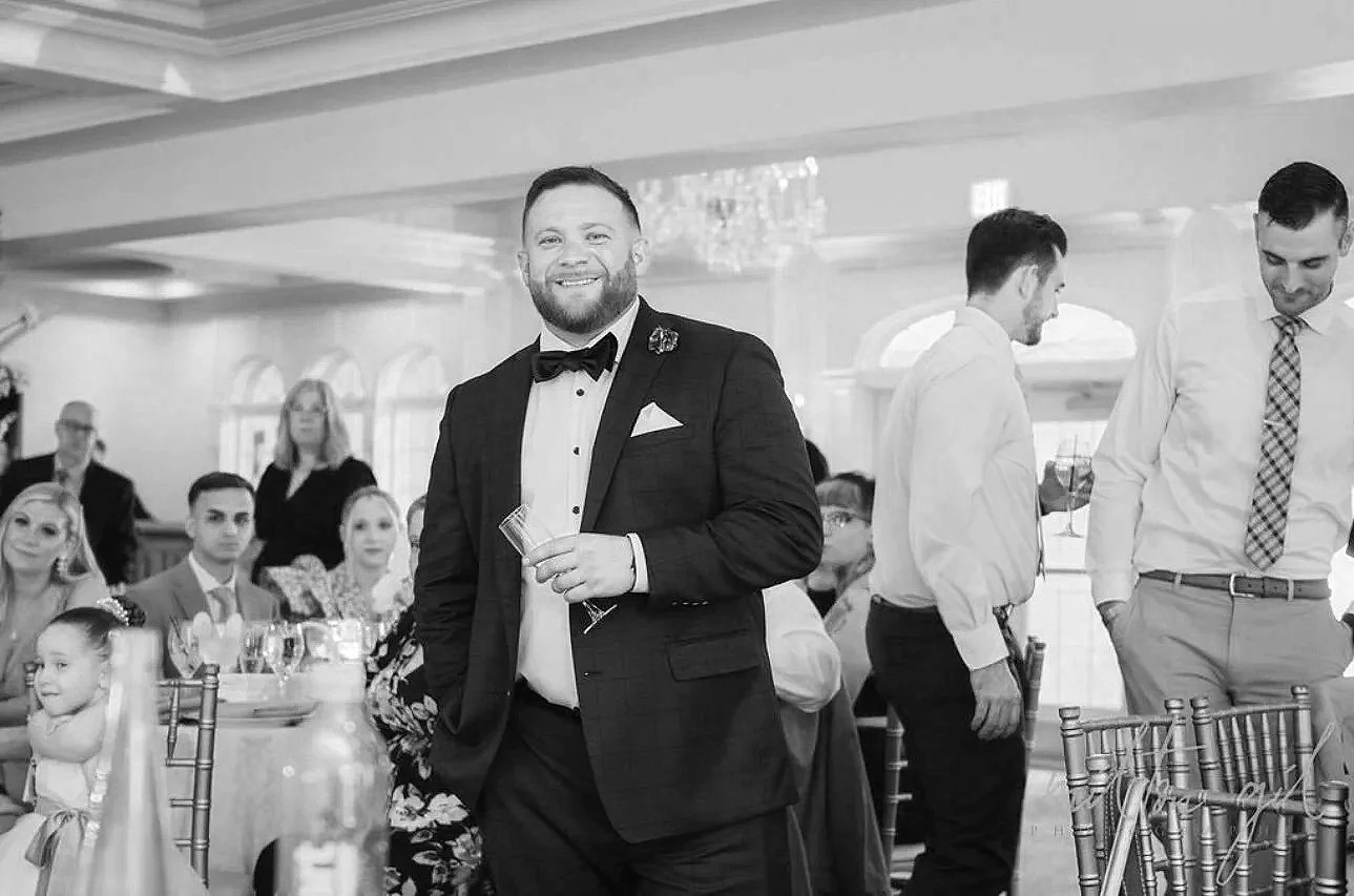 A man in a tuxedo holding a drink, smiling at a formal event with other guests seated at tables in the background.
