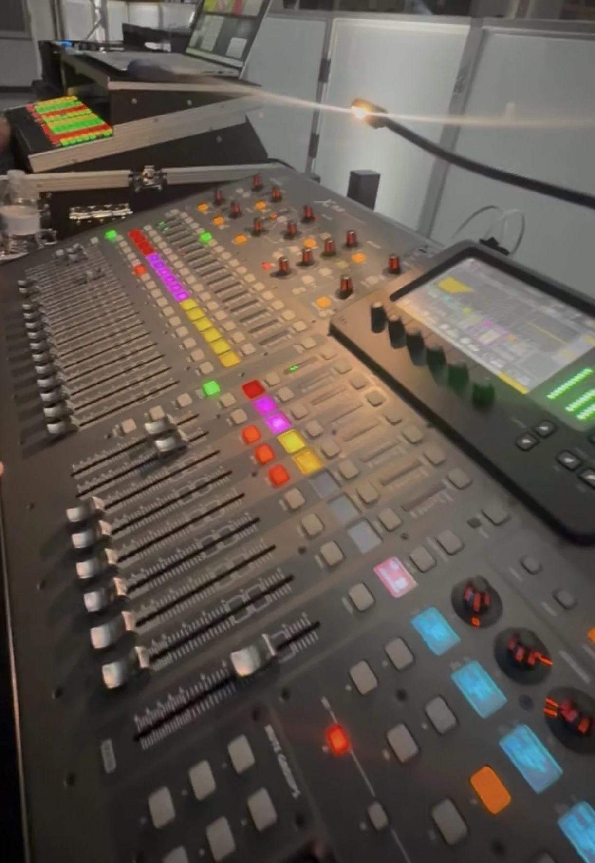 A professional audio mixing console with numerous sliders, knobs, and colorful buttons, along with a tablet device displaying audio levels and controls.
