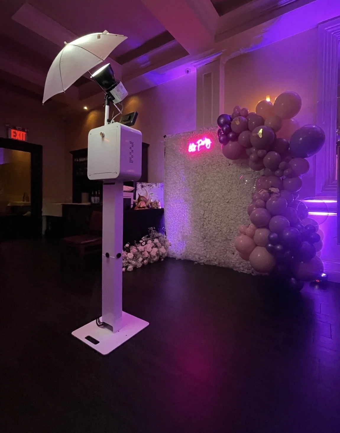 Event photo booth with balloon decorations, a pink neon sign, and a photo booth camera setup in a dimly lit room with purple lighting.