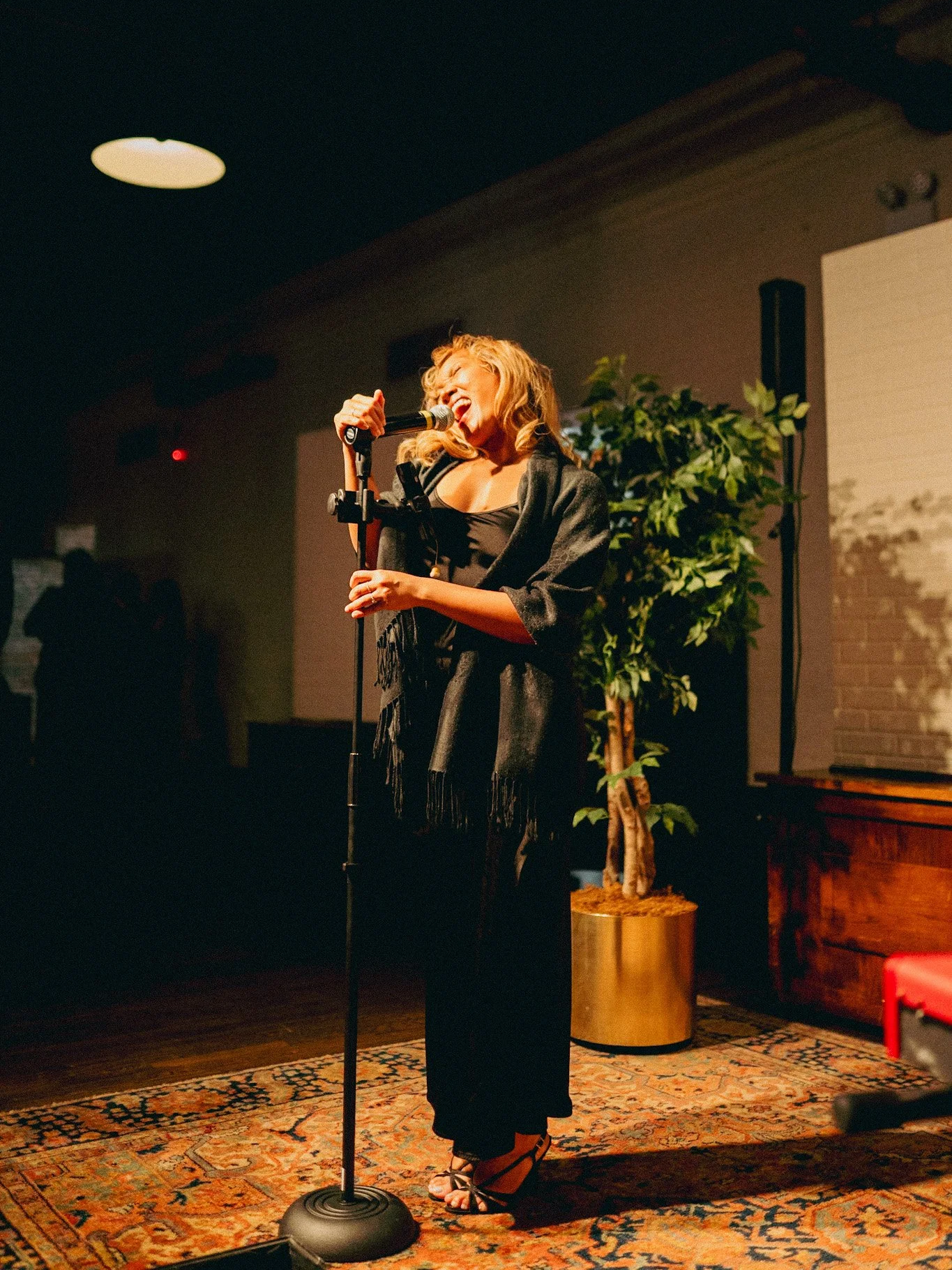 Woman singing on stage with microphone, standing on a patterned rug, in a dimly lit venue with plants and dark walls.
