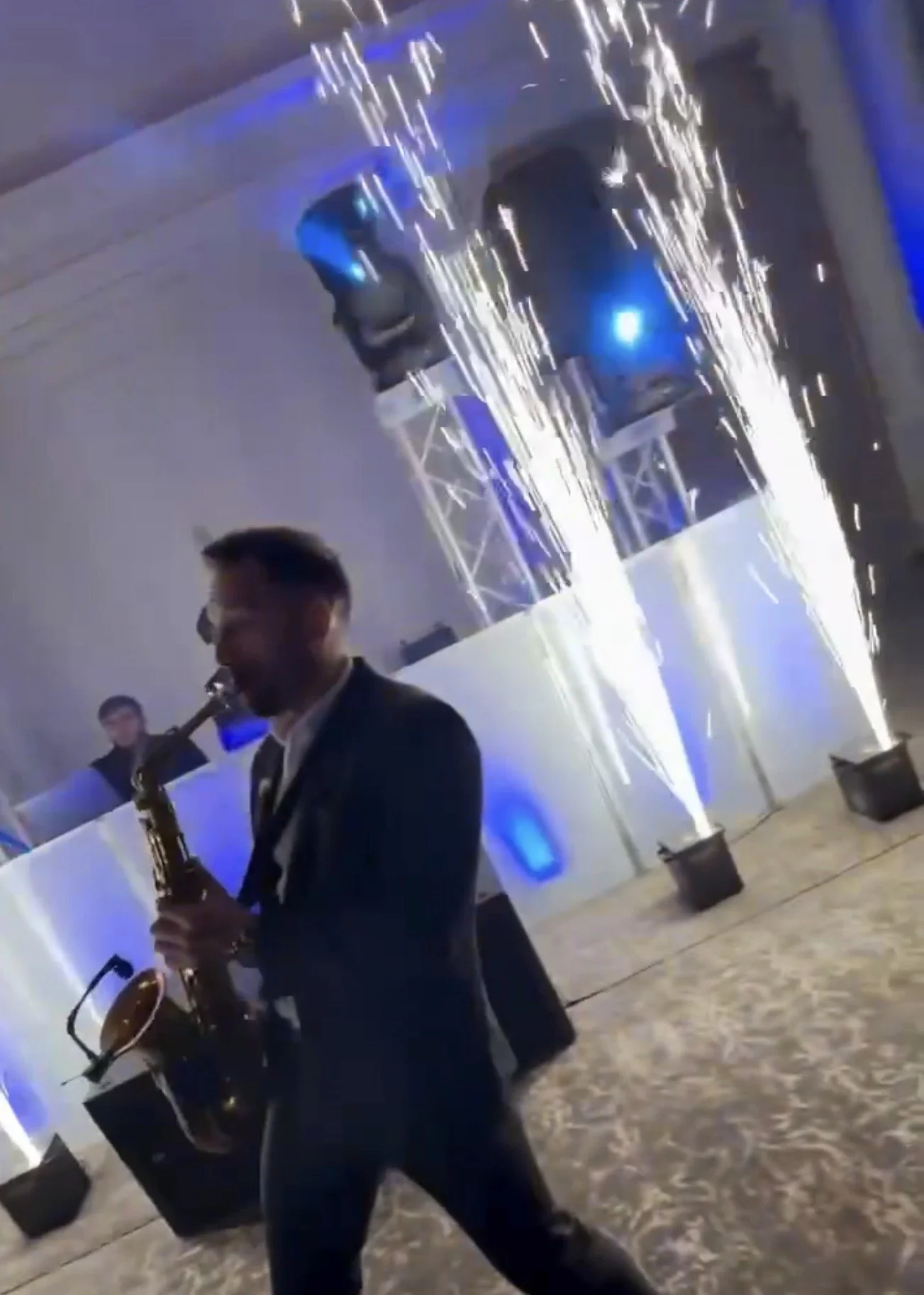 A man in a suit playing a saxophone at an indoor event with fireworks and blue lighting in the background.