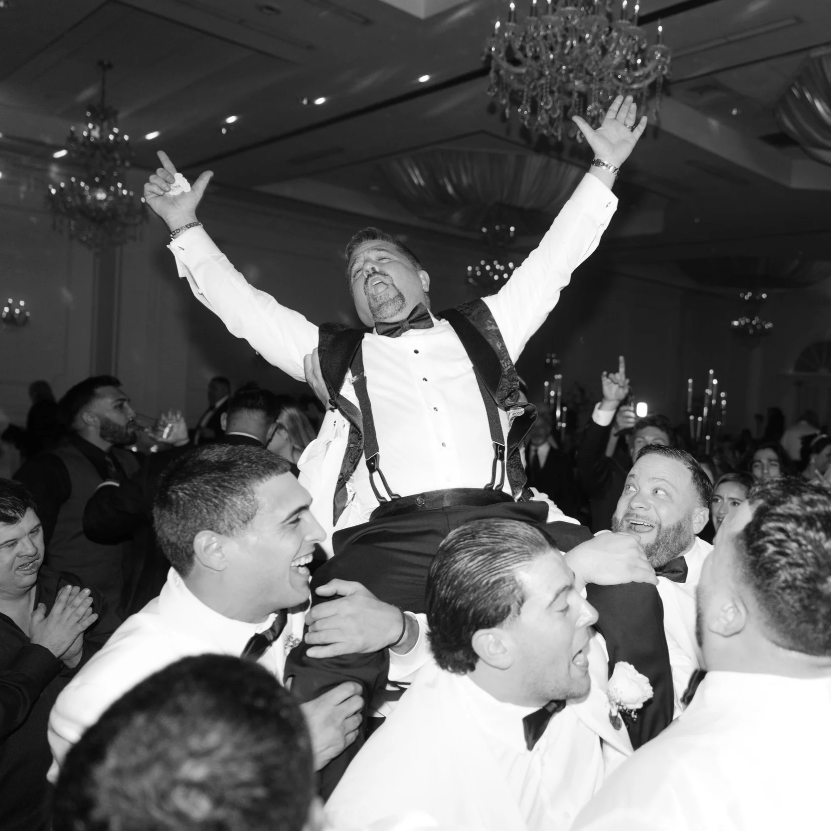 Group of men in tuxedos celebrating at a formal event with one man lifted up and raising his arms in excitement.