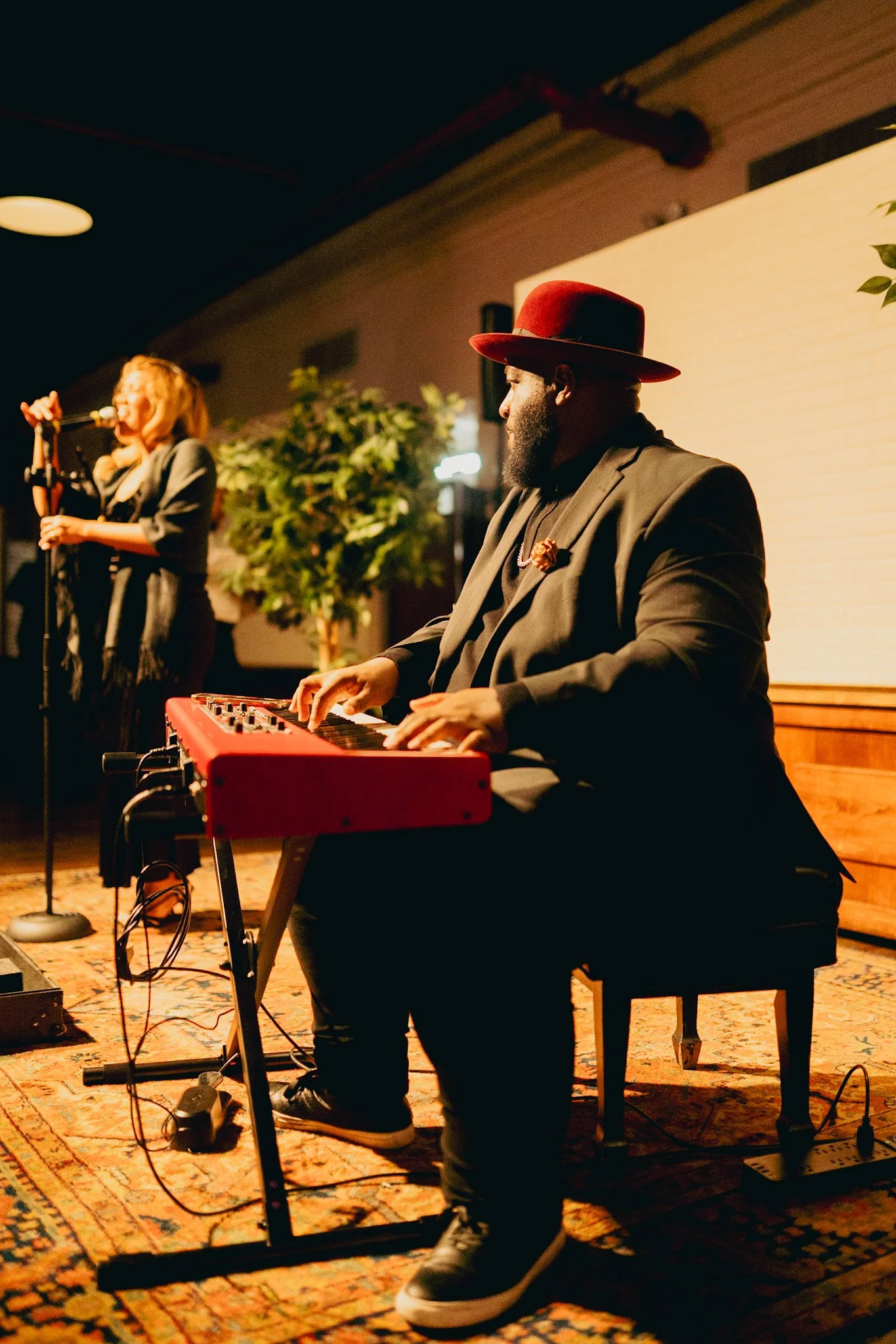A man wearing a red hat and black suit playing a red keyboard on stage, while a woman in the background sings into a microphone.