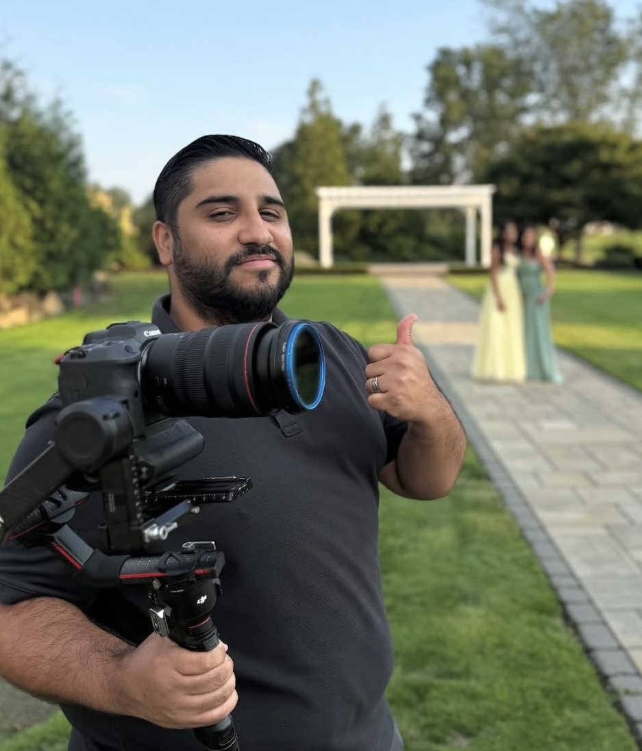 A man holding a professional camera on a stabilizer, giving a thumbs up at an outdoor wedding venue with two women in dresses in the background.
