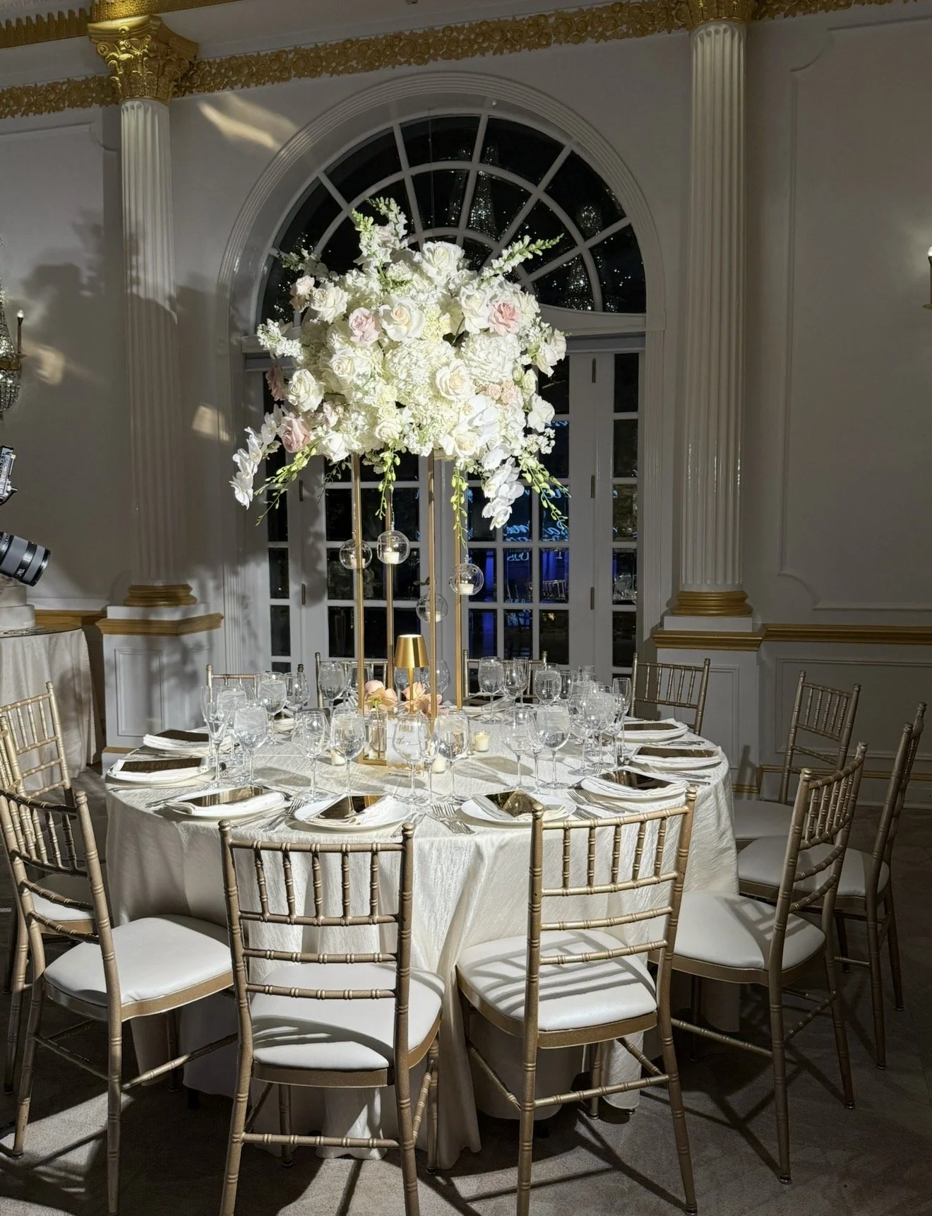Elegant round banquet table set for a formal event with white tablecloth, gold chairs, wine glasses, and floral centerpiece with white and pink flowers.