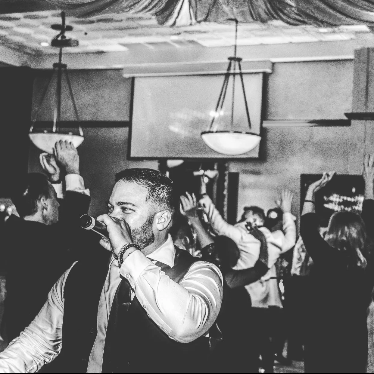 A man singing into a microphone while other people raise their hands in the background at a gathering or party.