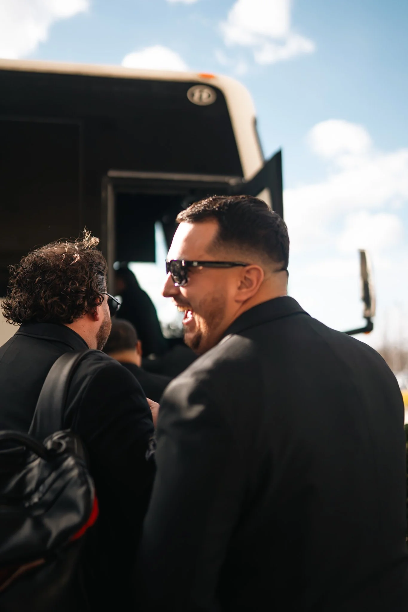 Two men in black suits and sunglasses talking near a large vehicle outdoors on a sunny day.