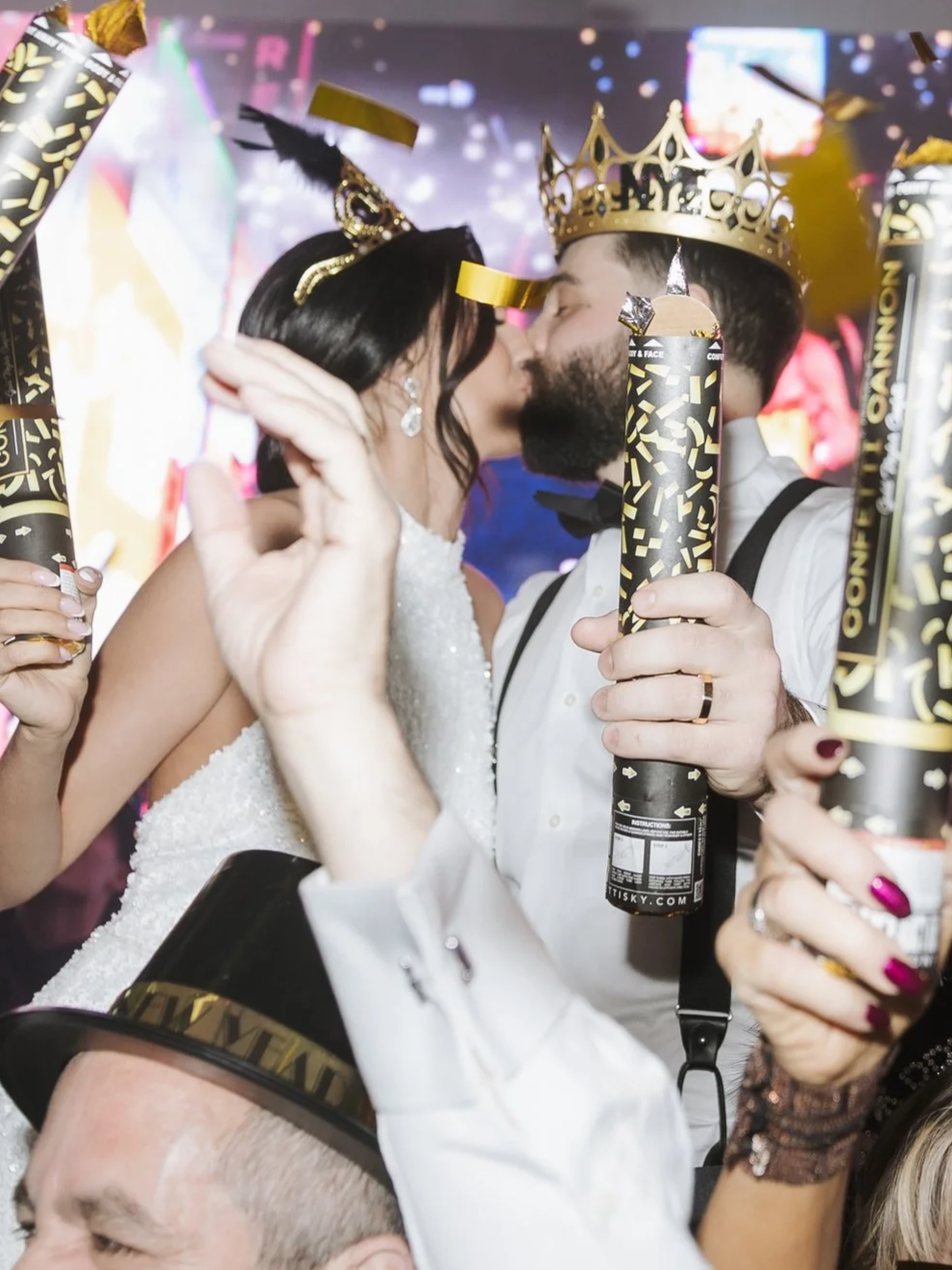 A couple dressed in wedding attire sharing a kiss at their celebration, with guests holding confetti cannons and wearing crowns and party hats.