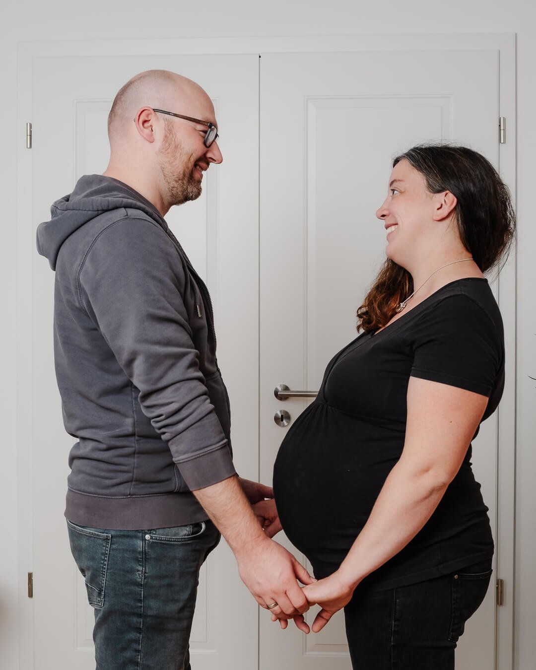eine schwangere Frau und ihr Mann stehen sich gegenüber, halten sich an den Händen und schauen sich lächelnd in die Augen bei einem Babybauch-Shooting bei Augsburg