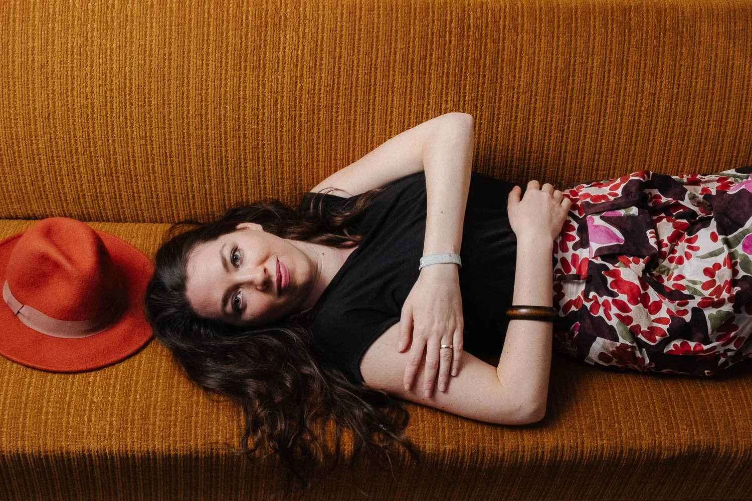 Portrait einer jungen Frau mit langen, braunen Haaren, einem schwarzen Top und einem bunten Rock, die auf einem Sofa im Stückwerk Krumbach neben ihrem roten Hut liegt und in die Kamera blickt