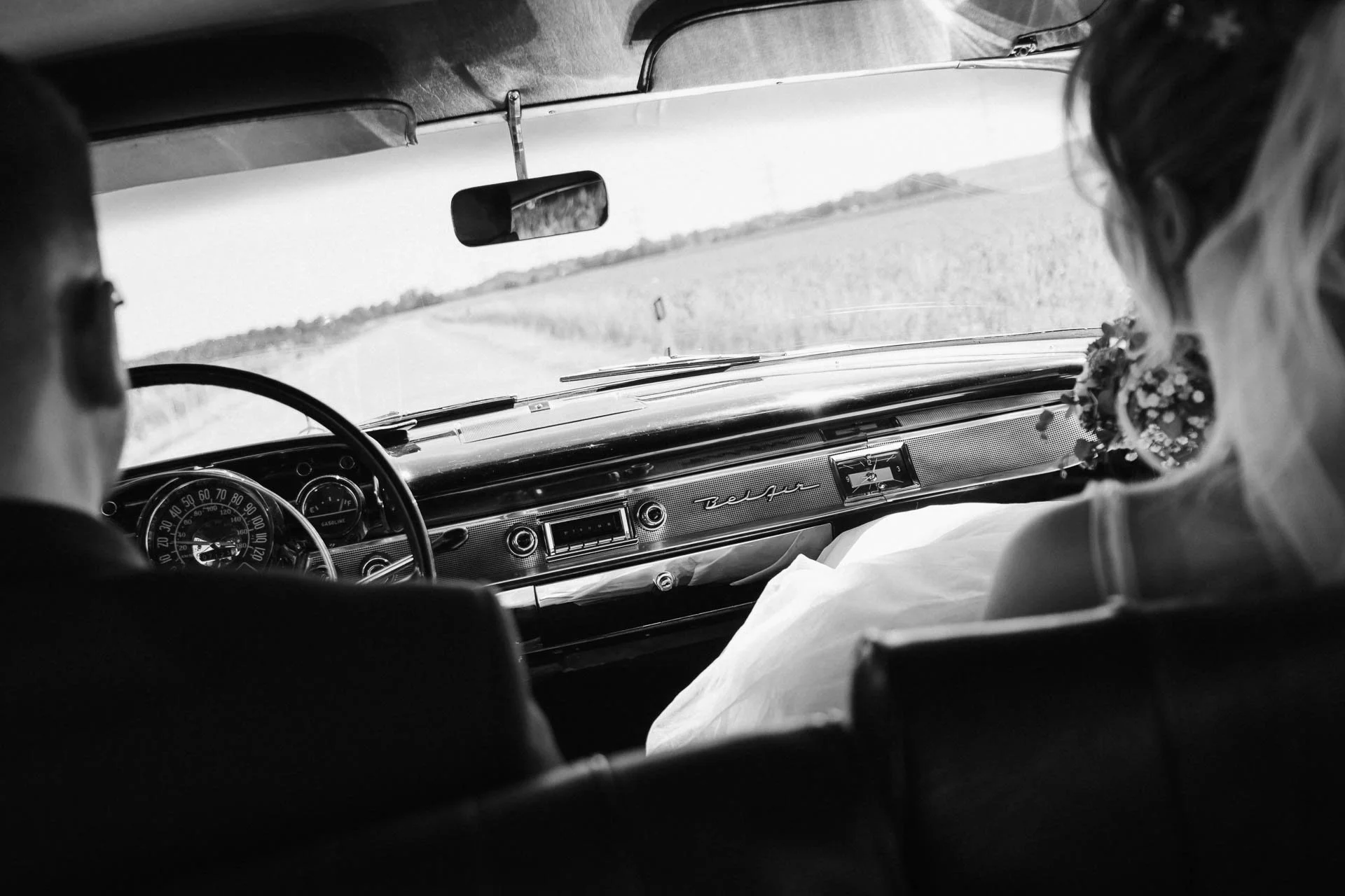 schwarz-weiß Foto, das ein Hochzeitsauto, einen Chevrolet-Oldtimer, von Innen zeigt und vom Rücksitz aus nach vorne aufgenommen ist, so dass man das Chockpit und das Brautpaar von hinten sieht