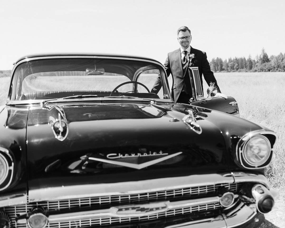 Schwarz-weiß Porträt eines Bräutigams, der an der Tür seines Hochzeitsautos, eines Oldtimers, steht und in die Kamera blickt, während im Hintergrund eine Wiese in der Umgebung von Augsburg zu sehen sind