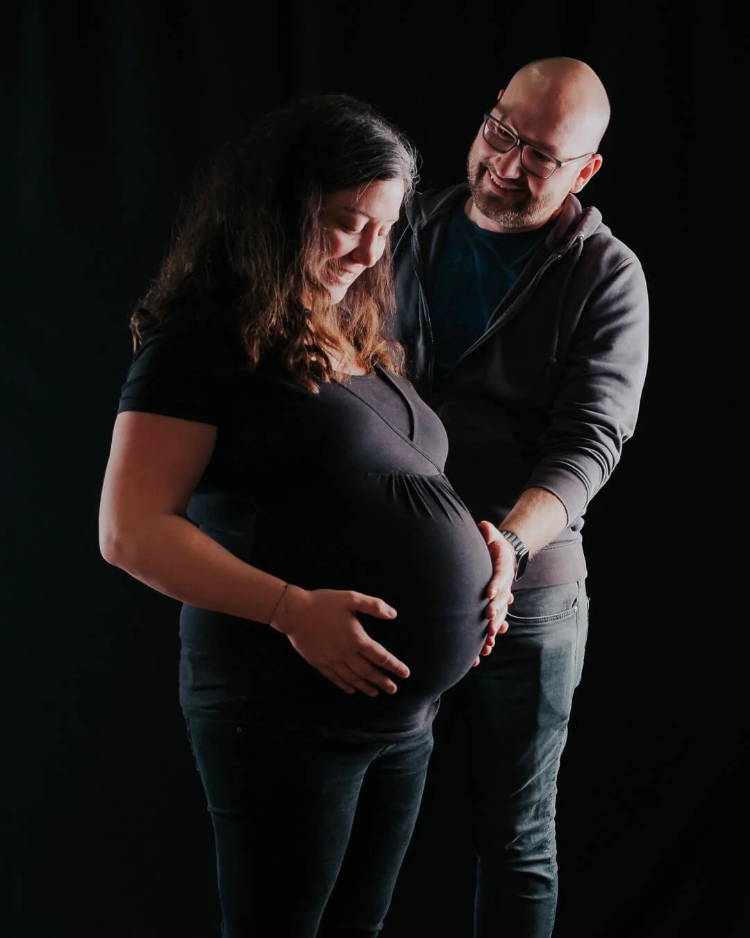 Paar mit schwangerer Frau beim professionellen Babybauch-Shooting im Fotostudio bei Augsburg mit schwarzem Hintergrund
